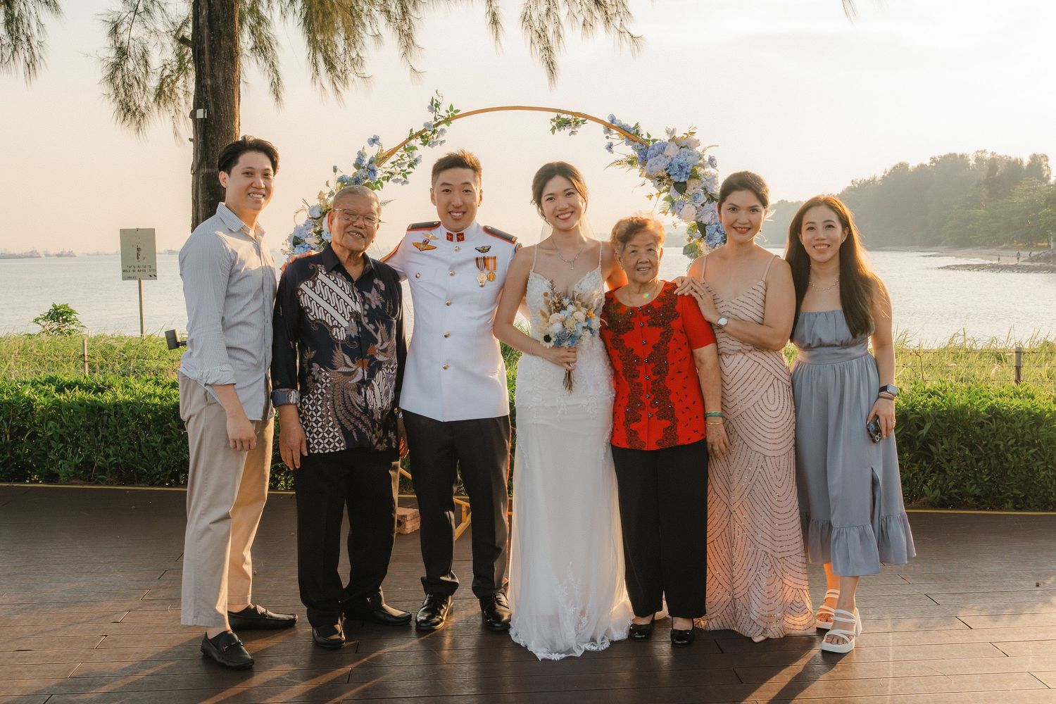Guests gathered for formal wedding photos at sunset near a floral arch and waterfront.