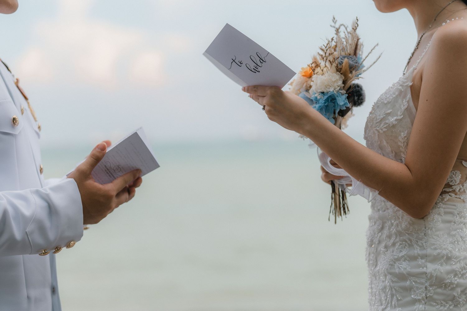 Couple reading vows at a seaside wedding ceremony with bouquet of blue and white flowers.