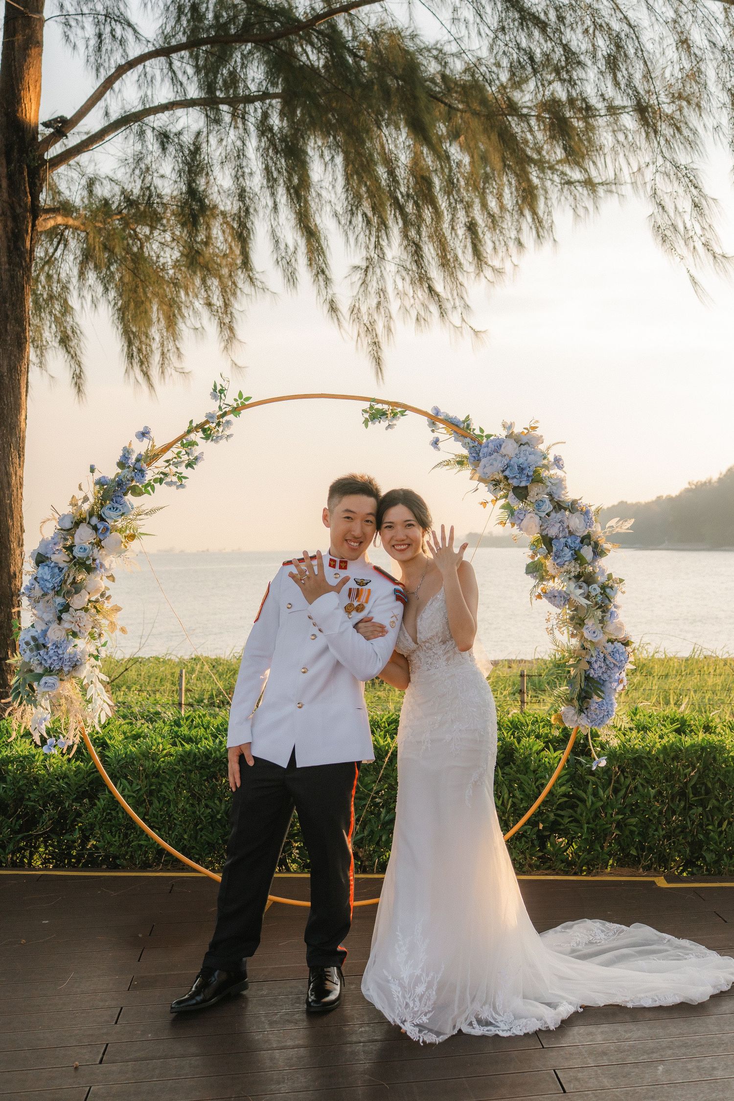 Wedding couple posing under a blue and white floral arch with ocean view at sunset.