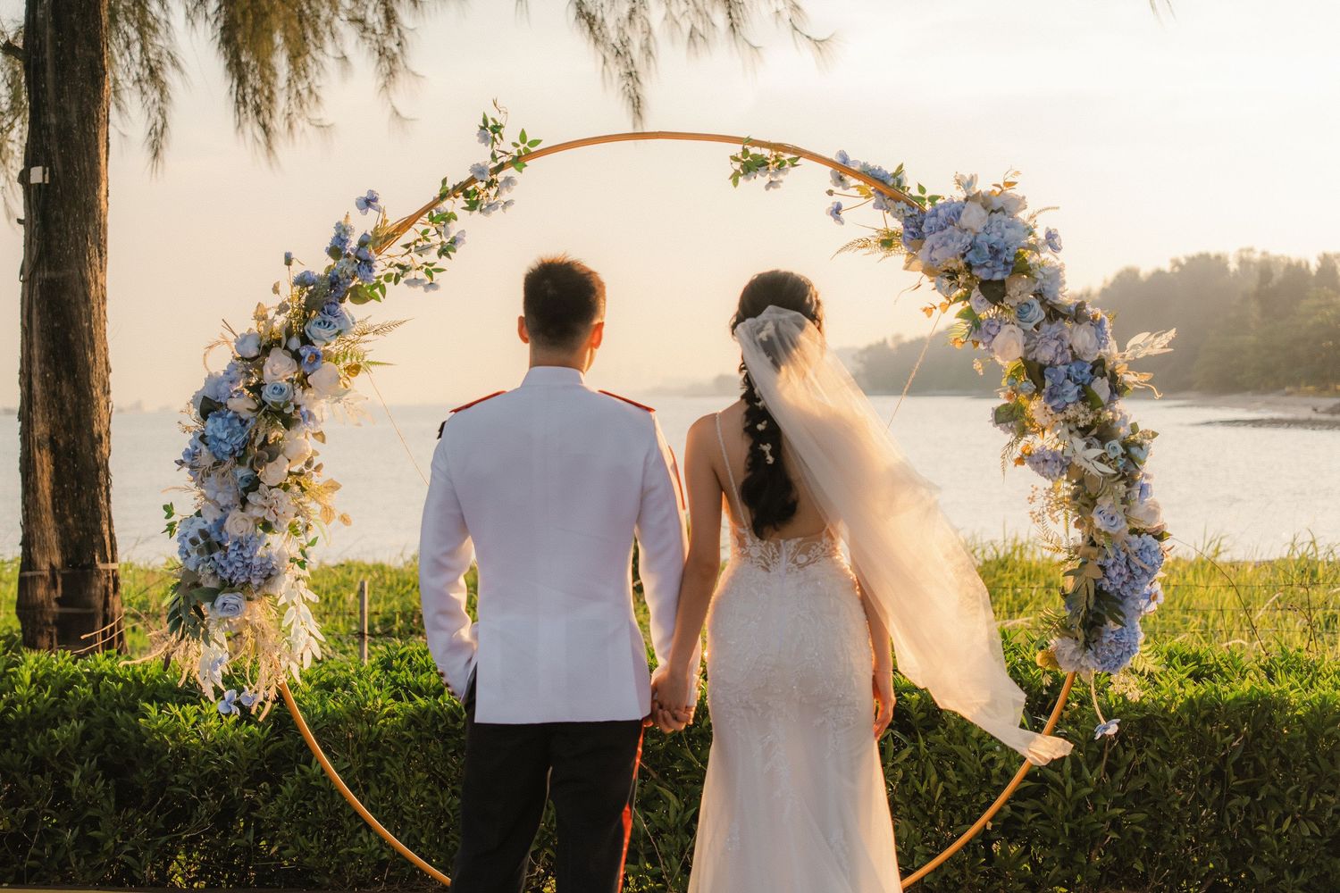 Silhouette of couple against sunset sky with floral wedding arch and waterfront view.