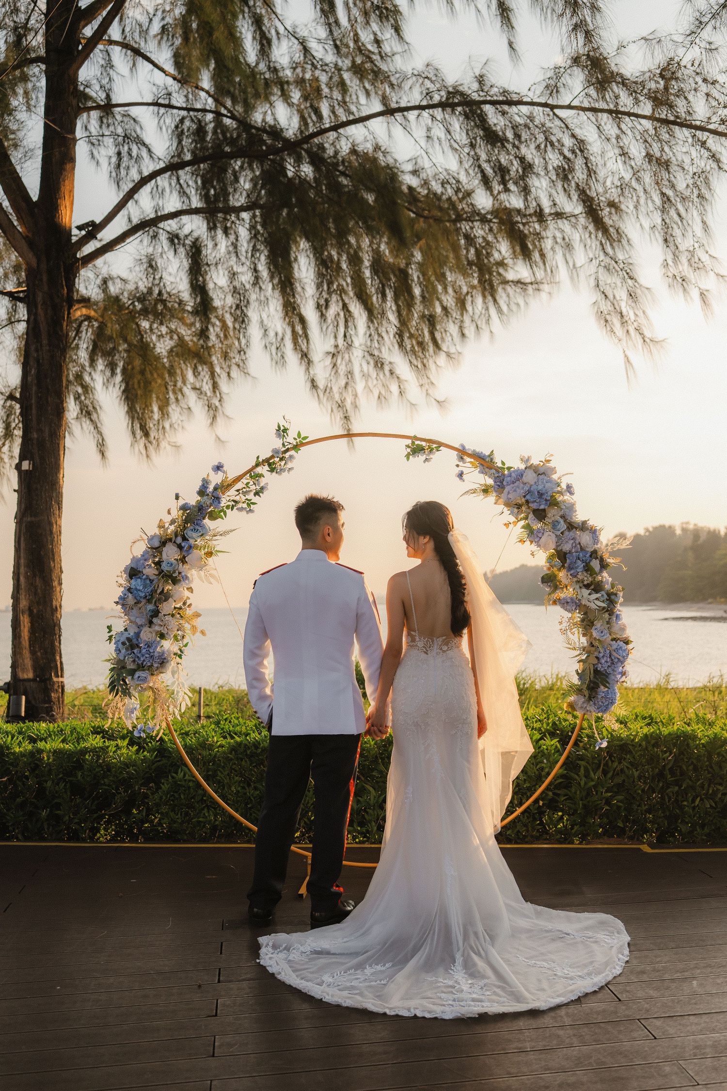 Elegant wedding couple walking along waterfront with floral arch and willow trees at sunset.