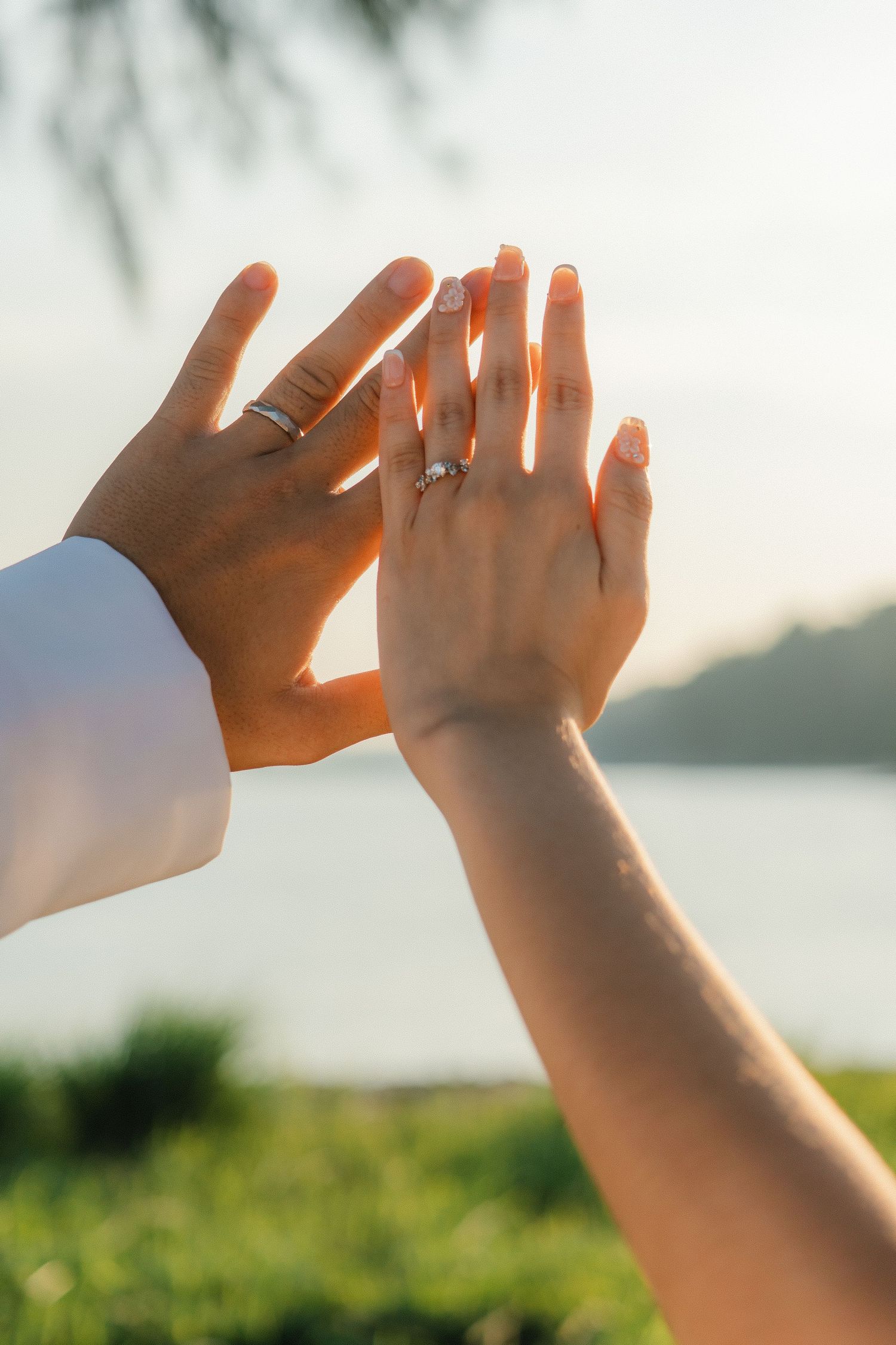 Two hands with wedding rings held up against a sunset waterfront backdrop.