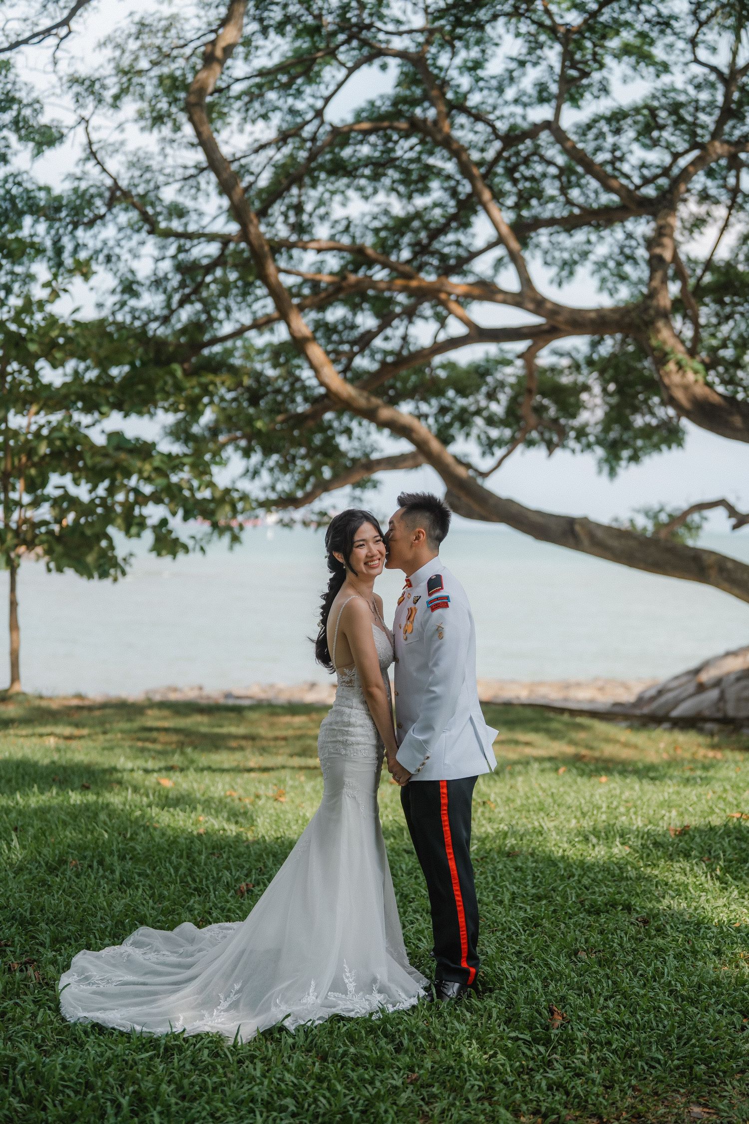 A military wedding couple poses under a large tree branch by the water in formal attire on a grassy lawn.