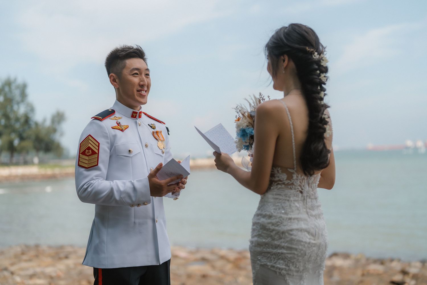 A couple in military dress uniform and wedding attire embracing on a beach with ocean views.