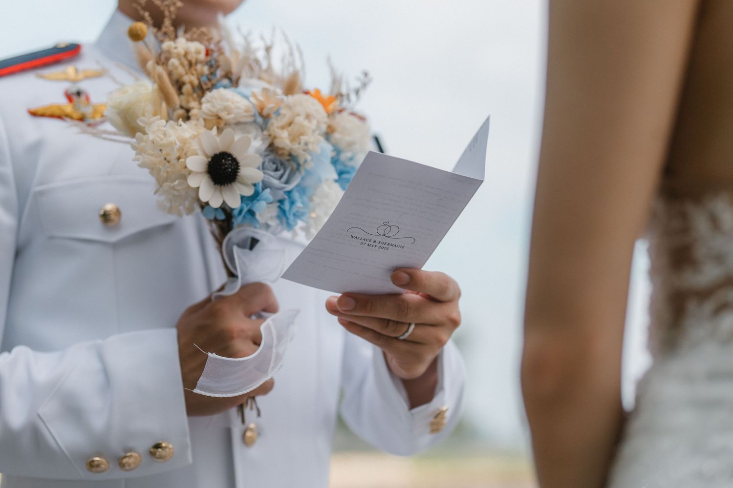 Someone in a white uniform holds a card while carrying a bouquet with dried flowers and blue accents during a wedding ceremony.