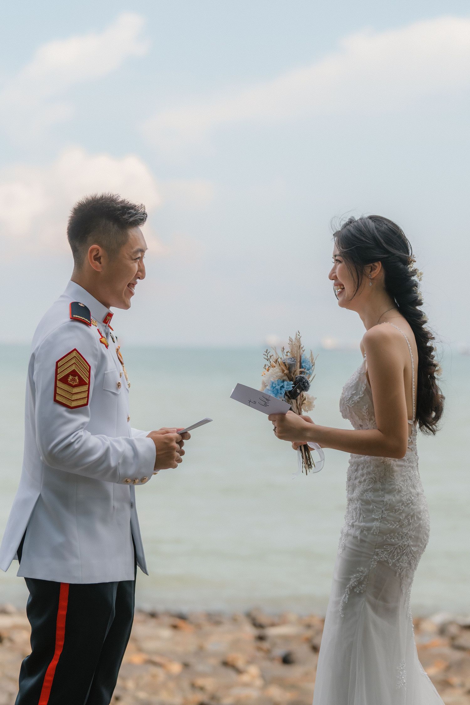 A couple in wedding attire exchange words during their ceremony by the seaside on a rocky shore.