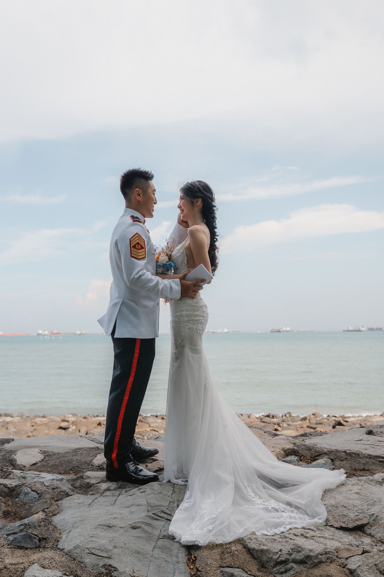A bride in a flowing white gown and groom in formal military uniform embrace on rocky coastline during sunset ceremony.