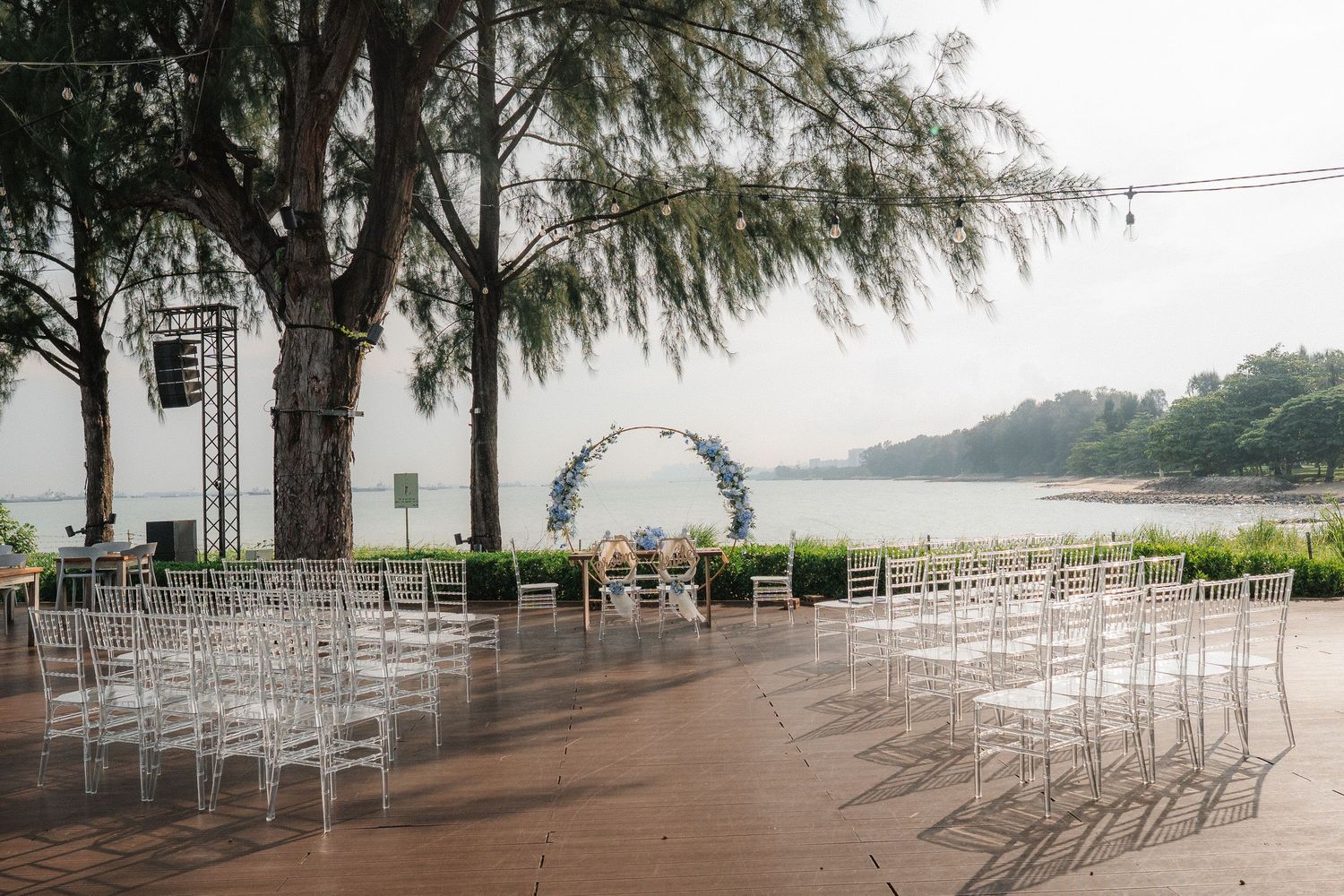 Empty white wedding chairs arranged outdoors on a terrace with an arch and tree overlooking a scenic water view.