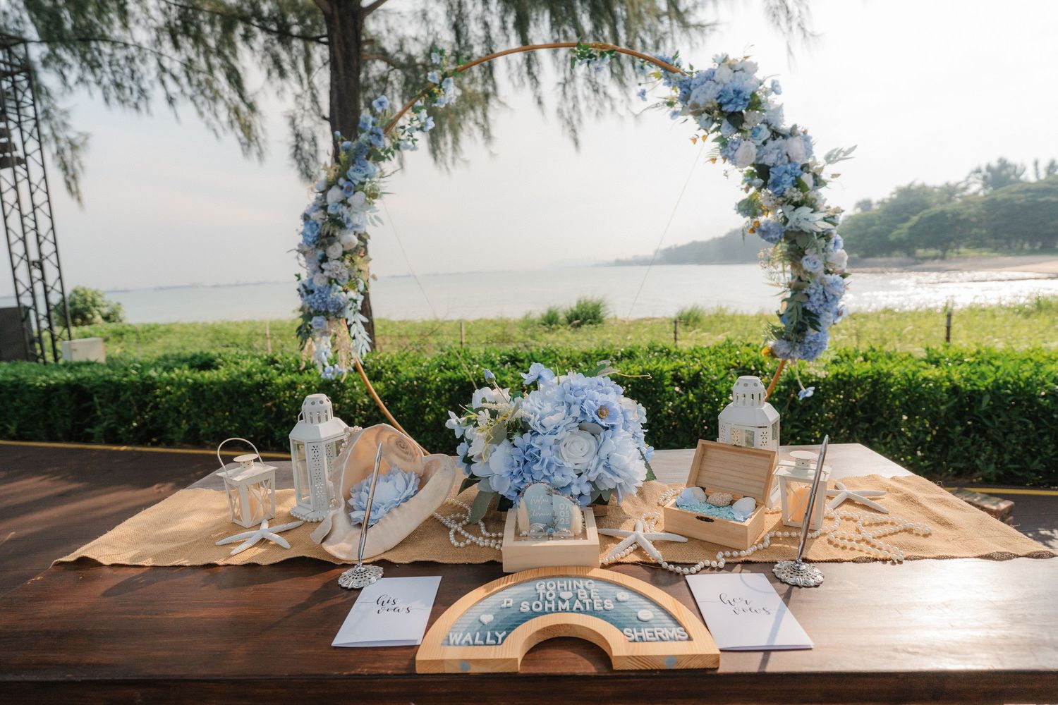 Elegant rustic wedding reception decor with blue floral arrangements and candles on a wooden table beneath pine trees.