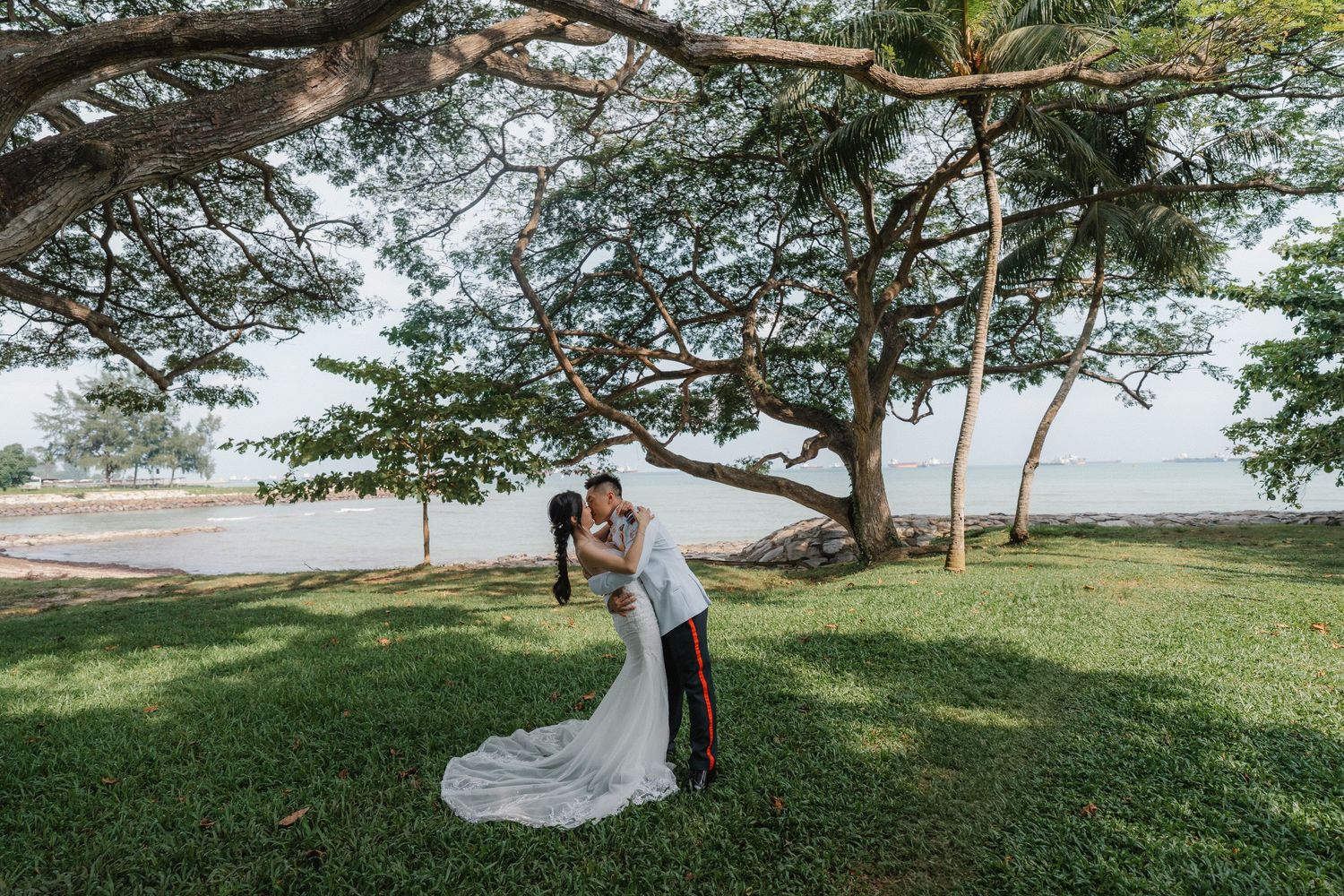 A couple shares a romantic embrace during their outdoor wedding photoshoot under lush green trees by the ocean.