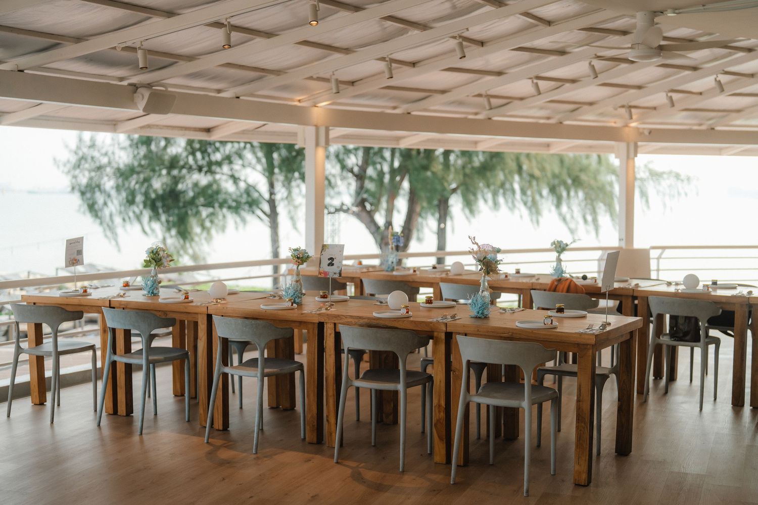 Empty beachfront restaurant interior with wooden tables and chairs arranged under a covered patio area overlooking the ocean.