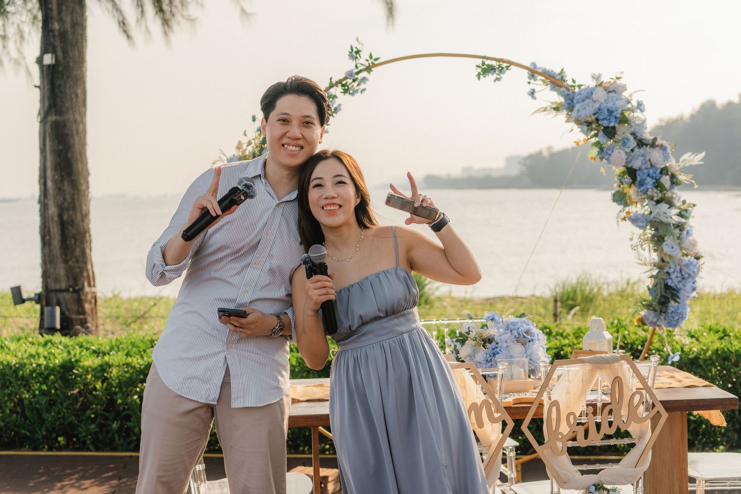 A joyful celebration under a floral arch with blue flowers by the waterfront at sunset with wooden chairs and decor.