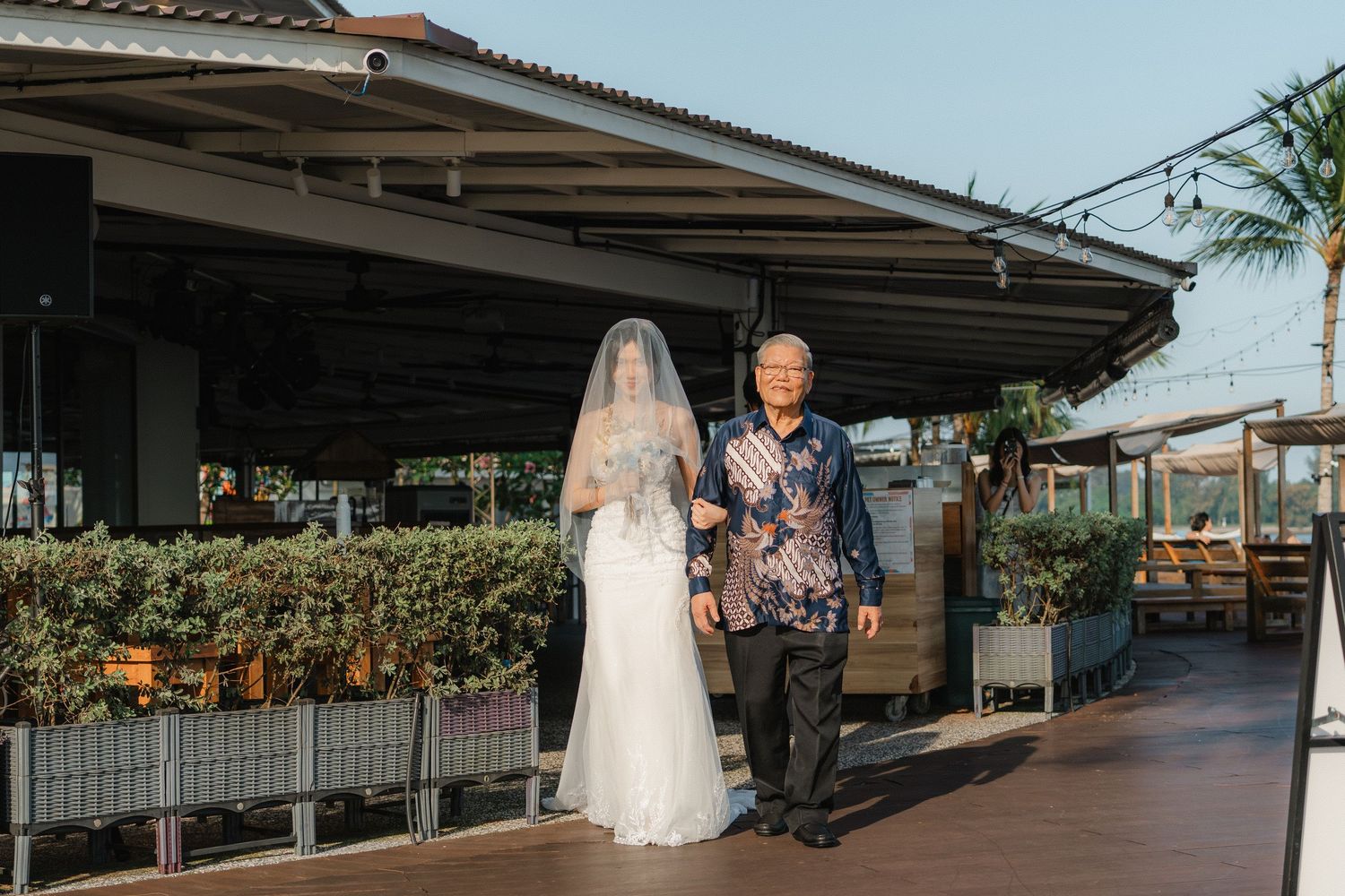A wedding ceremony takes place at a covered outdoor restaurant patio with tropical decor.
