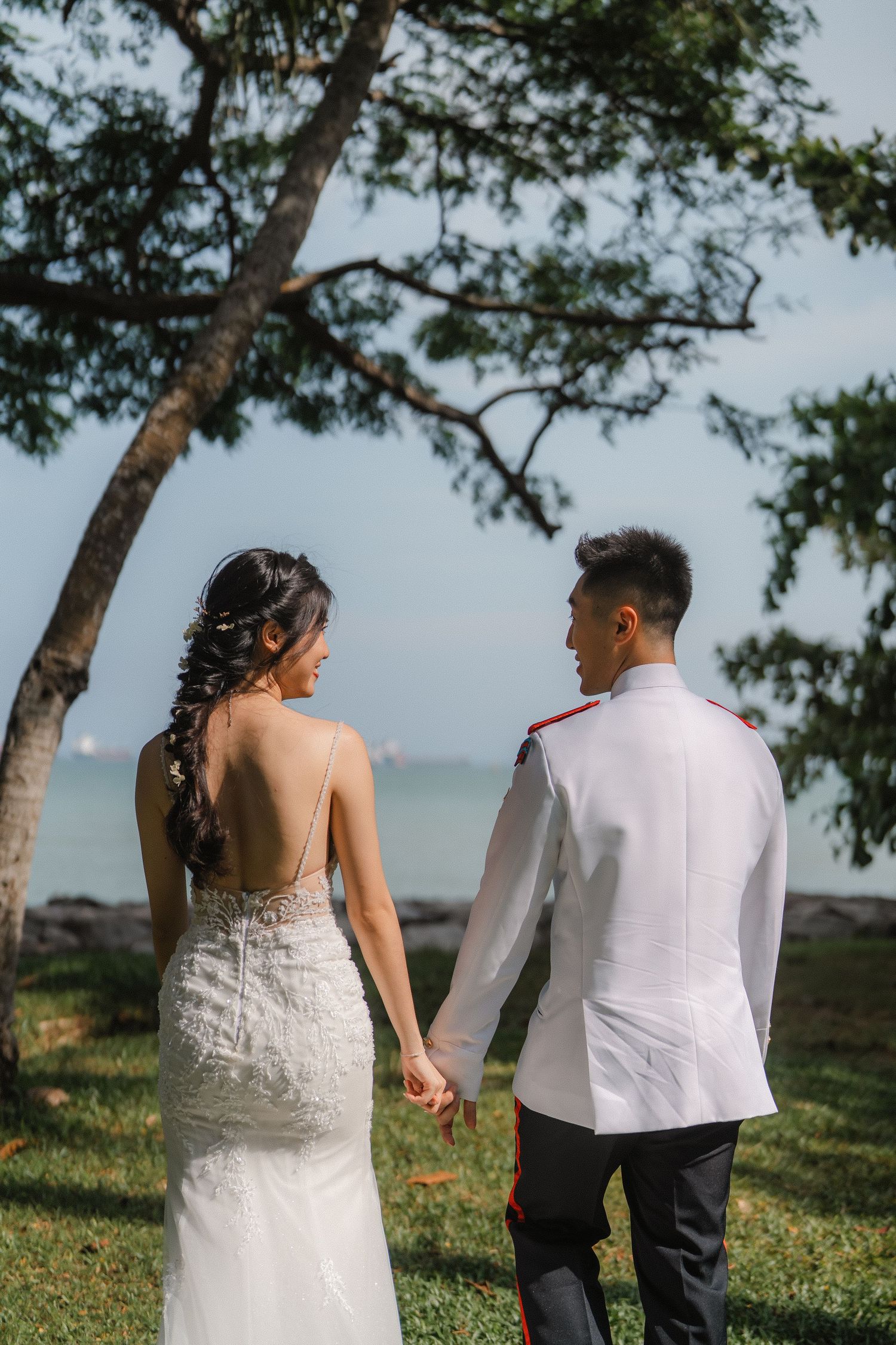 A wedding couple holds hands while walking on grass under a tree with ocean views in the background.
