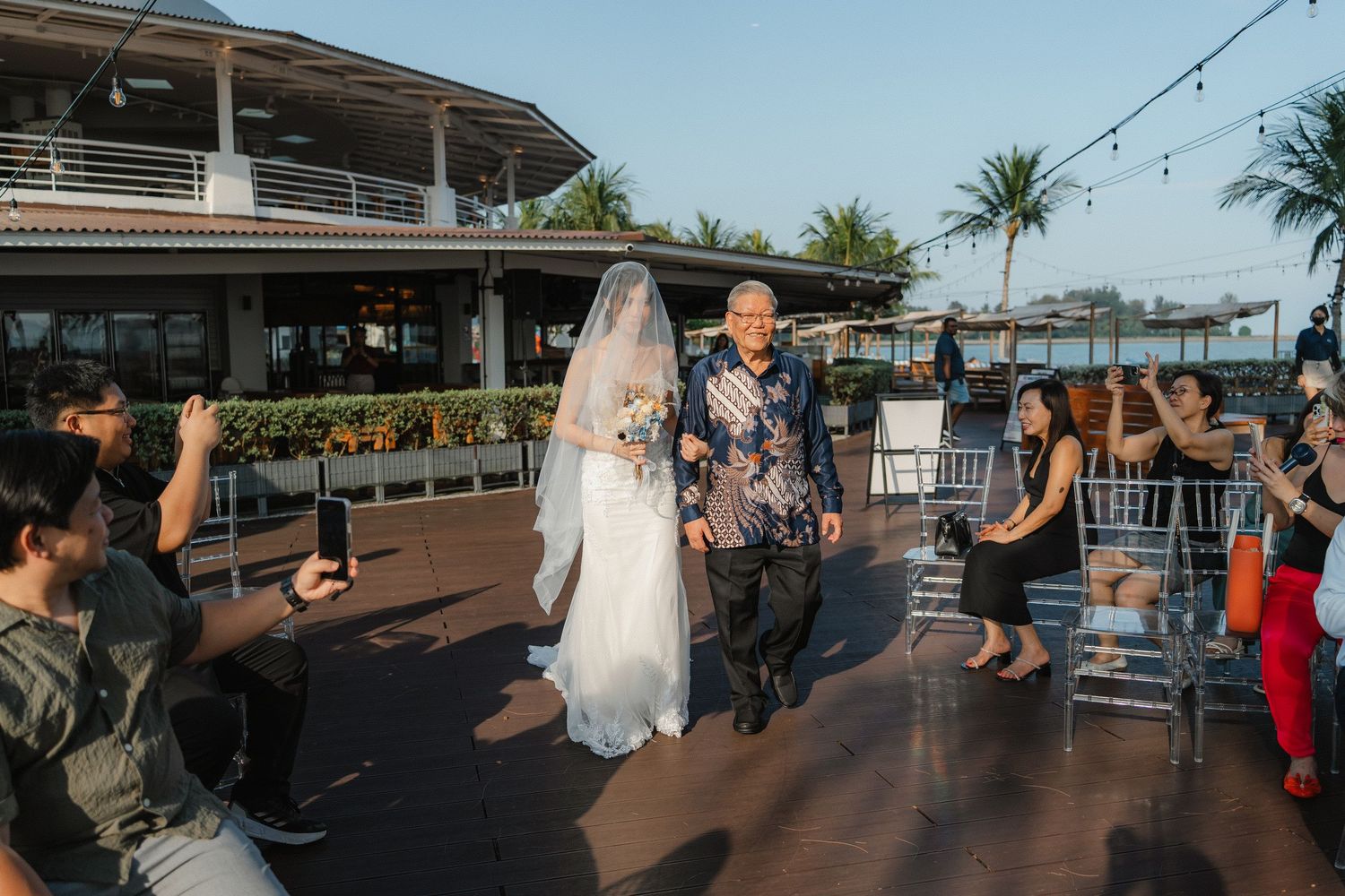 Wedding guests mingle on an outdoor waterfront deck with string lights overhead during twilight.