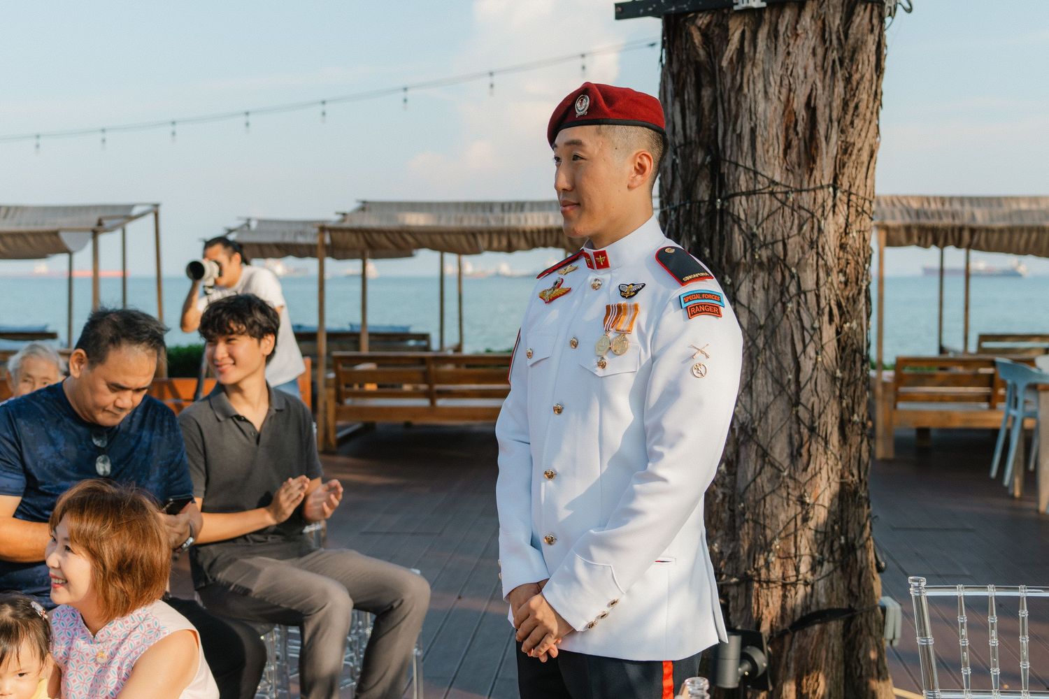 A military service member in dress whites stands at an outdoor venue with wooden benches and ocean views in the background.