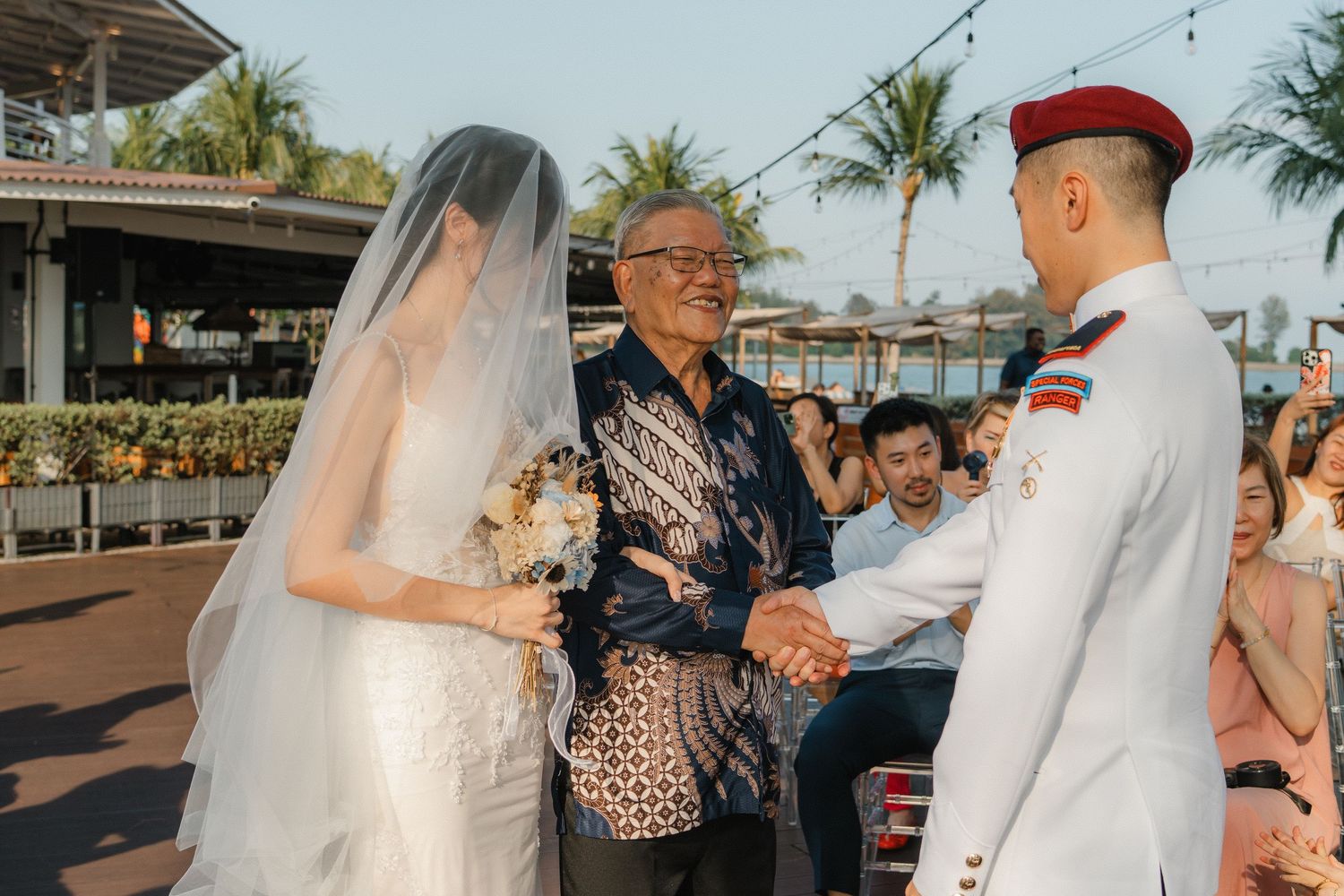A wedding ceremony takes place outdoors with palm trees in the background at a tropical venue.