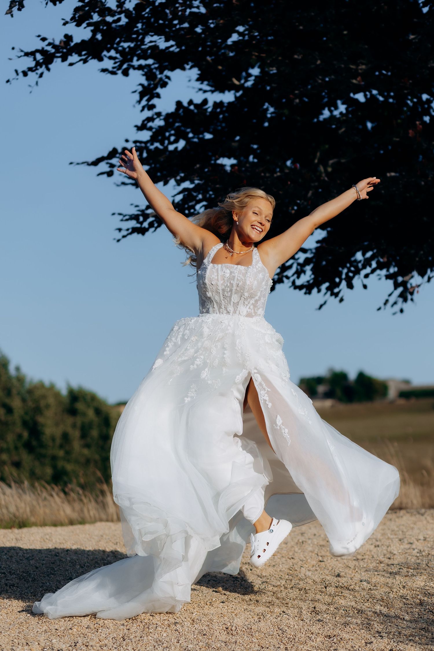 Joyful person in flowing white wedding dress twirls and dances outdoors on a sunny day in a rustic field setting.