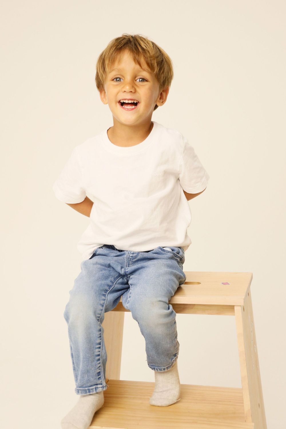 A child wearing a white t-shirt and jeans sits on a light wooden chair against a neutral background.