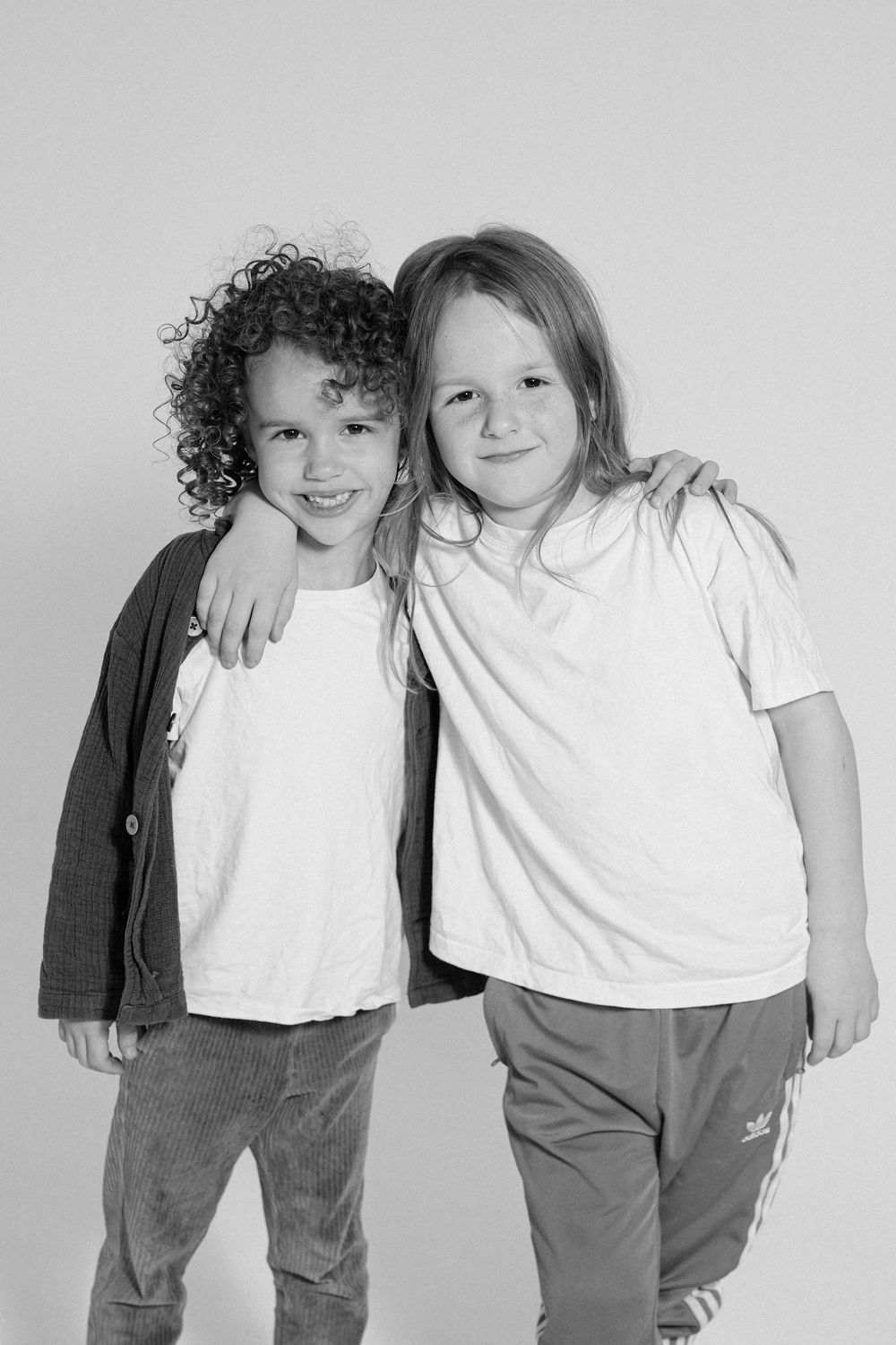 Two children in casual attire pose with arms around each other in monochrome studio shot