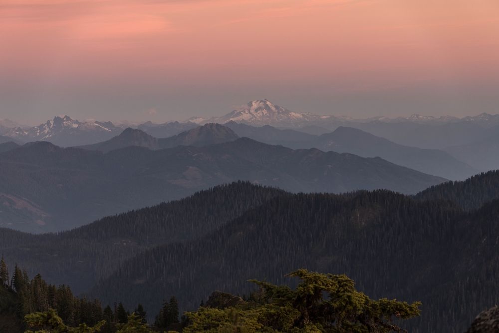 Scenic mountain range silhouettes at sunset with layered peaks fading into the distance under a pink and purple sky.