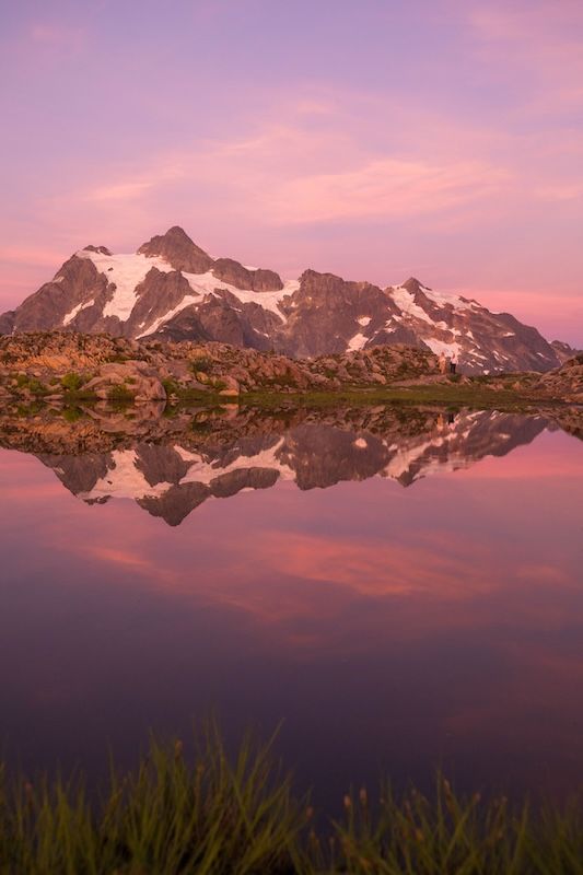Snowy mountain peaks reflect perfectly in a calm alpine lake during a pink sunset creating a mirror-like panorama.