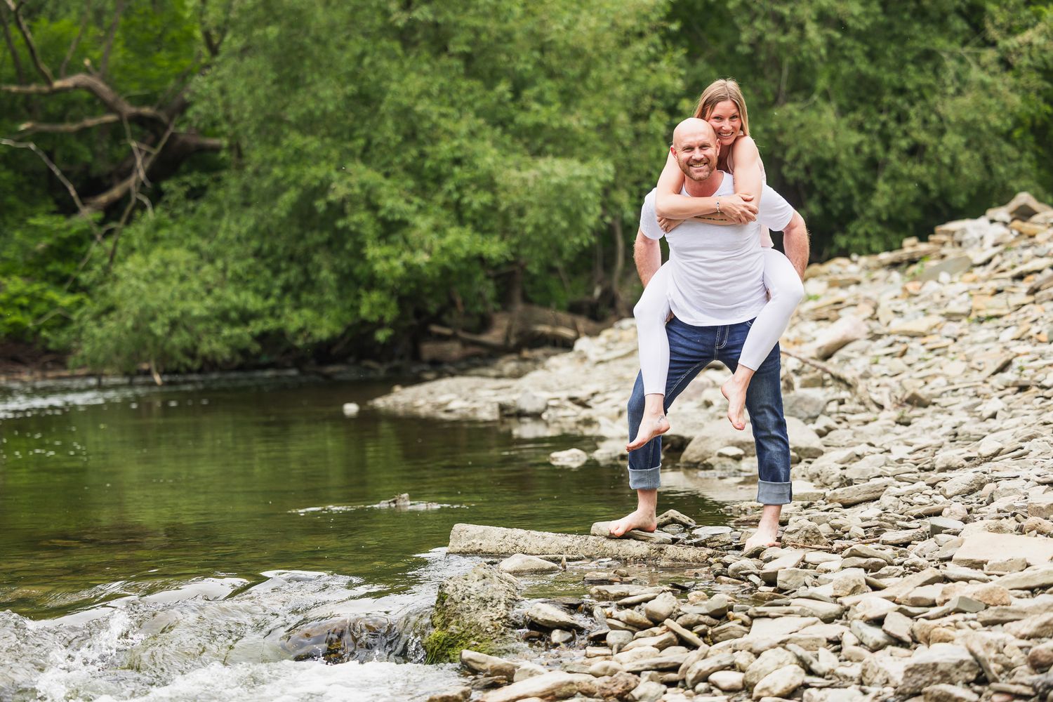 Two people having fun and laughing while balancing on rocks by a river.