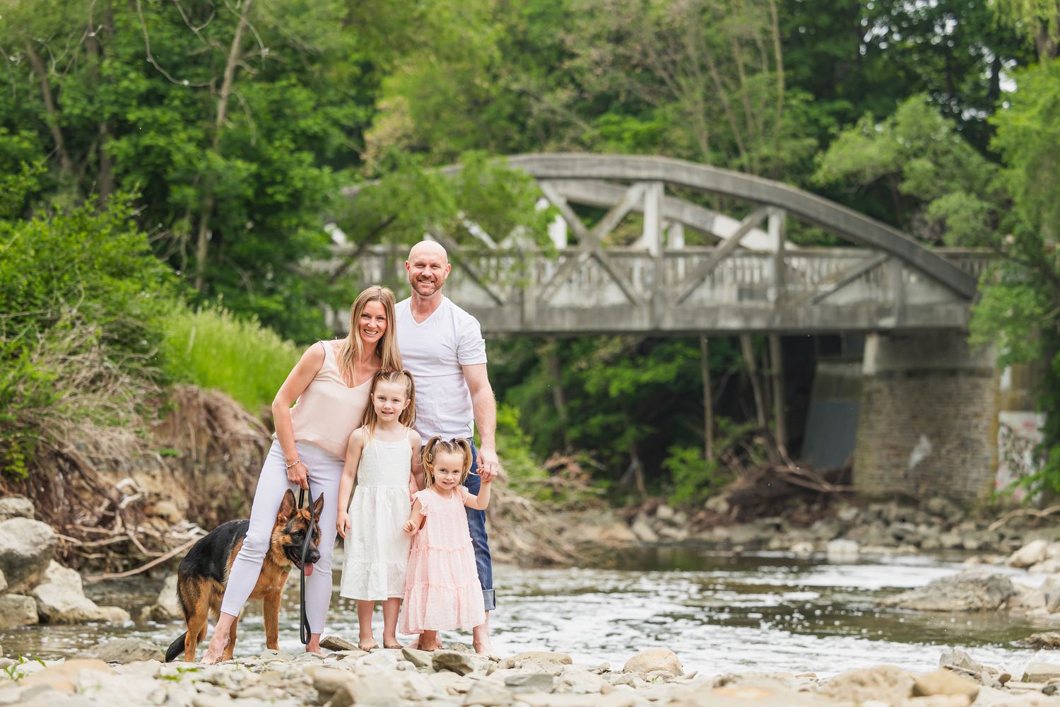 A family poses together on rocky riverbank with rustic bridge in background.