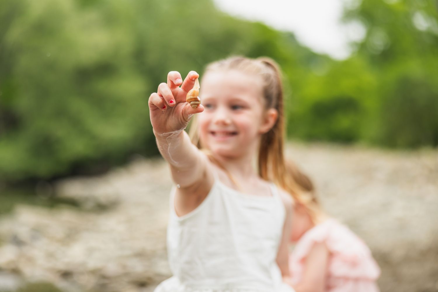 Child in white dress holding up something small while standing outdoors in soft focus.