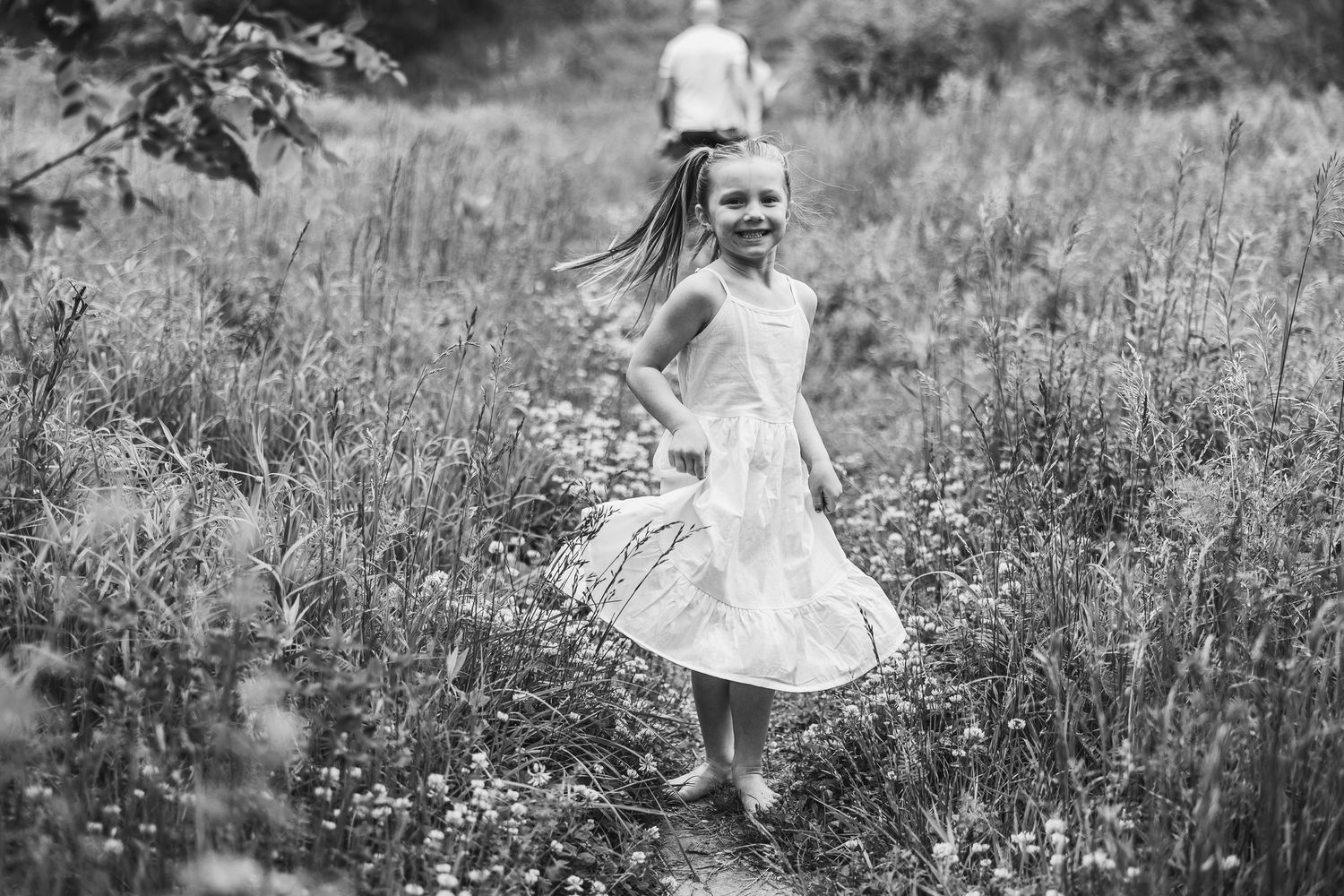 Child in a white dress twirling through a meadow of tall grass in black and white.