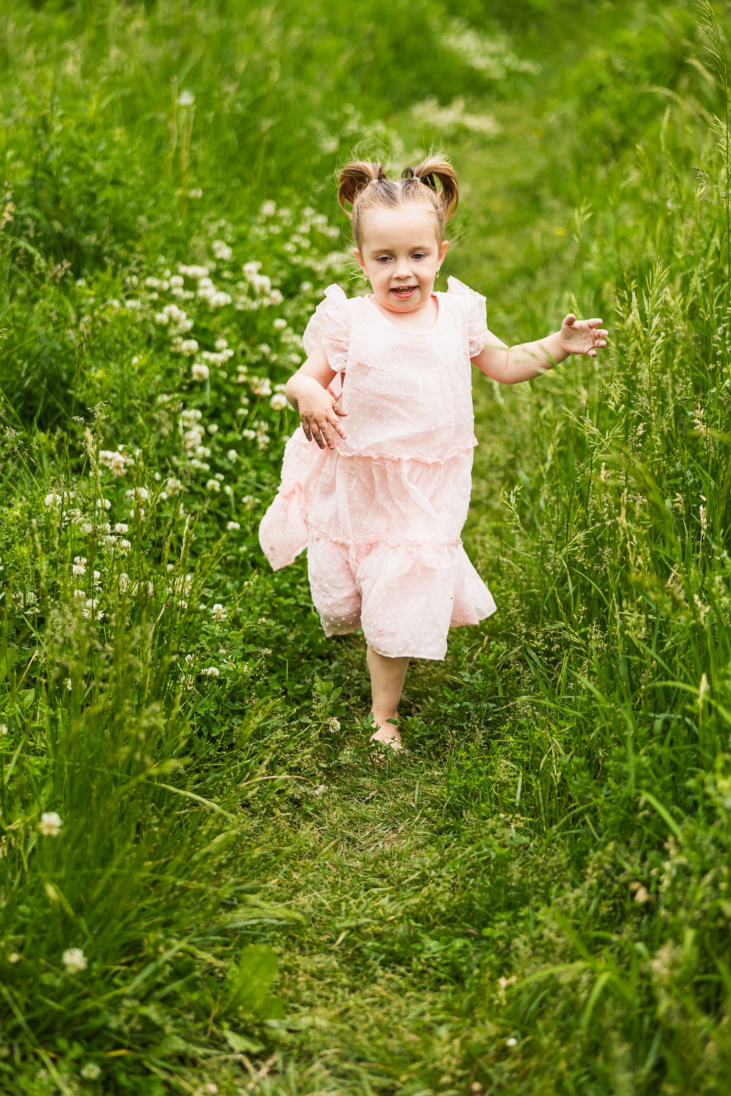 A small child in a pink dress runs joyfully through a meadow filled with wildflowers on a sunny day.