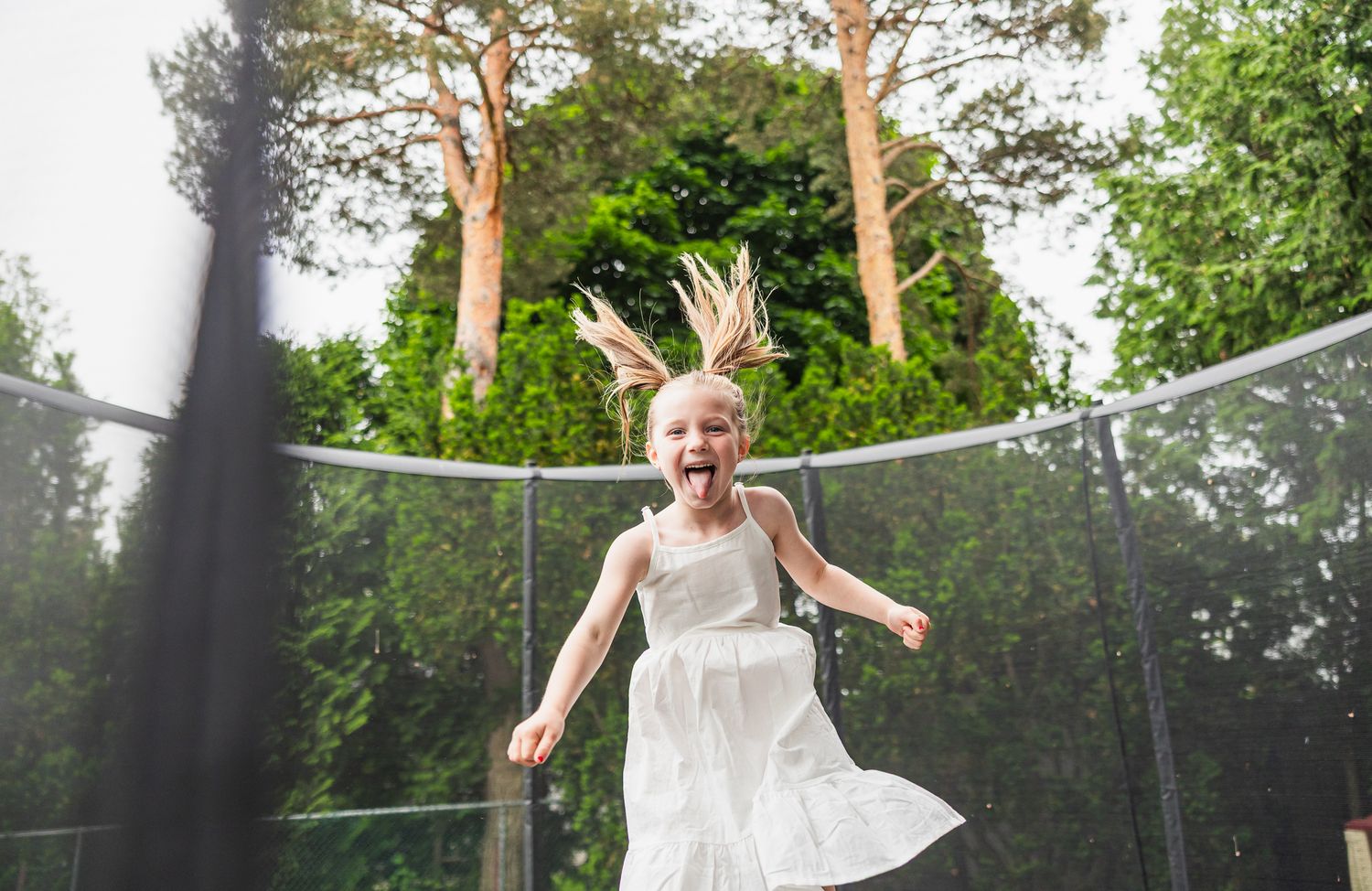 A child in a white dress jumps happily on an outdoor trampoline surrounded by tall trees.