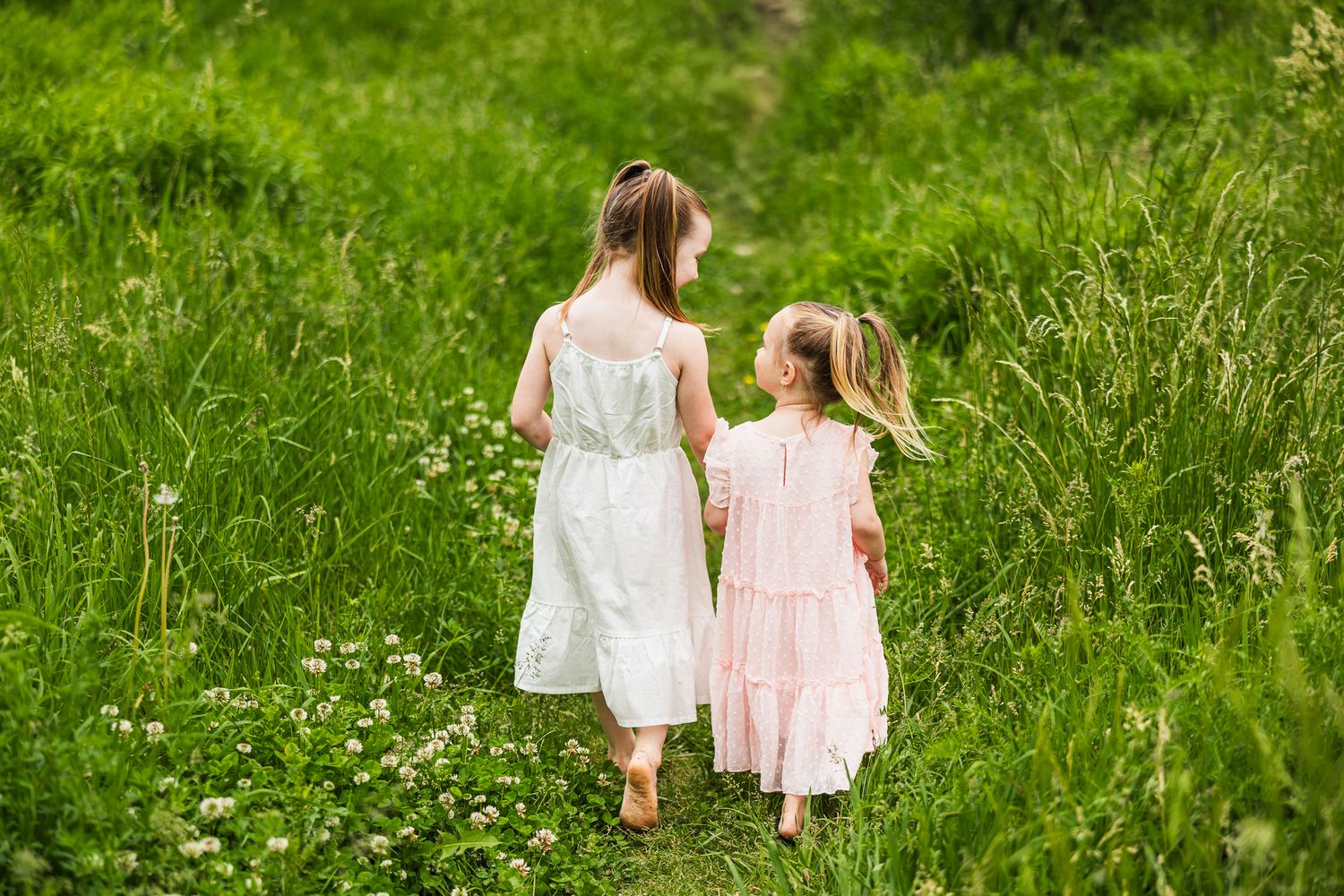 Two people in white and pink dresses walk together through a sunny meadow filled with wildflowers.