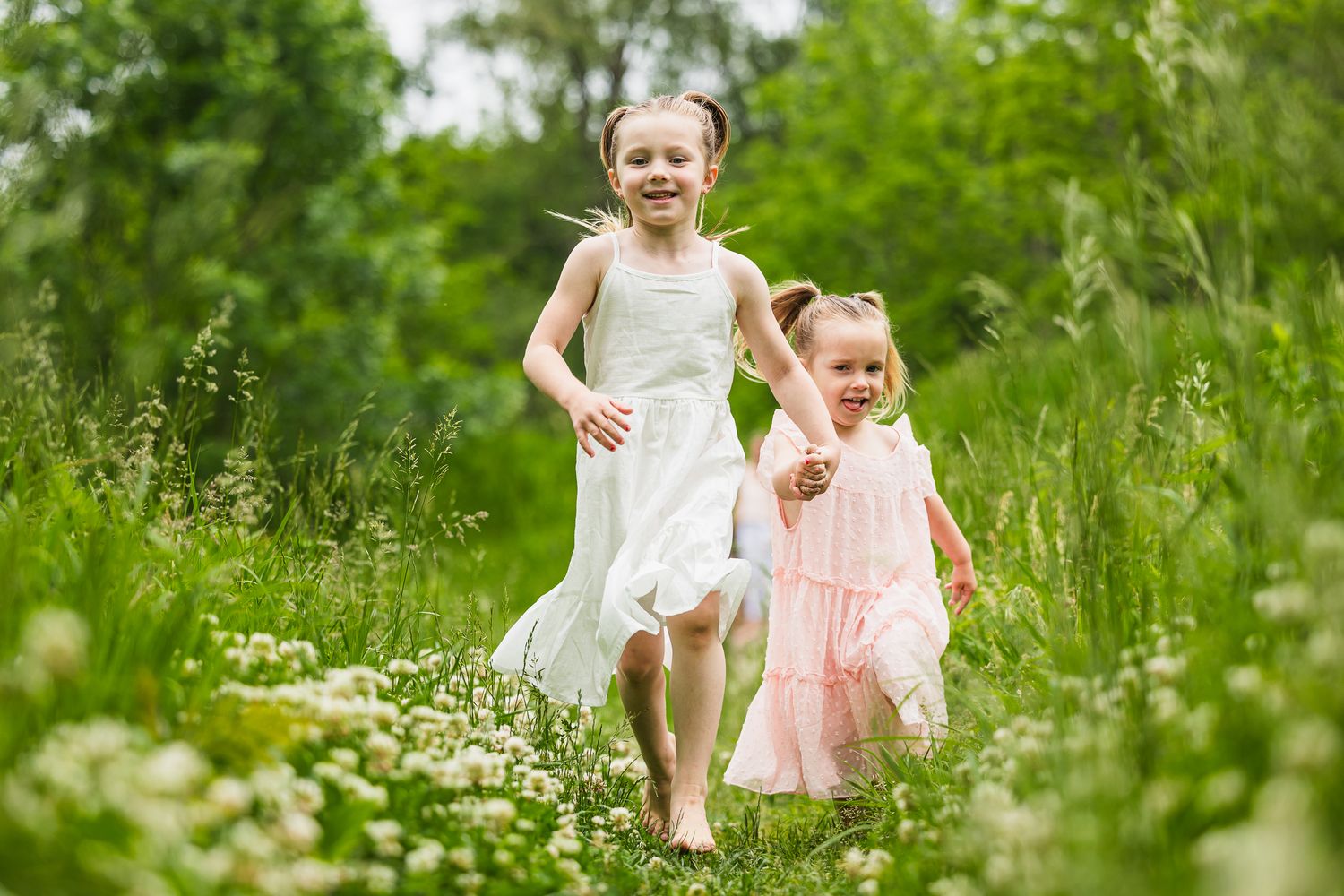 Two children in white and pink dresses running joyfully through a wildflower meadow on a sunny summer day.