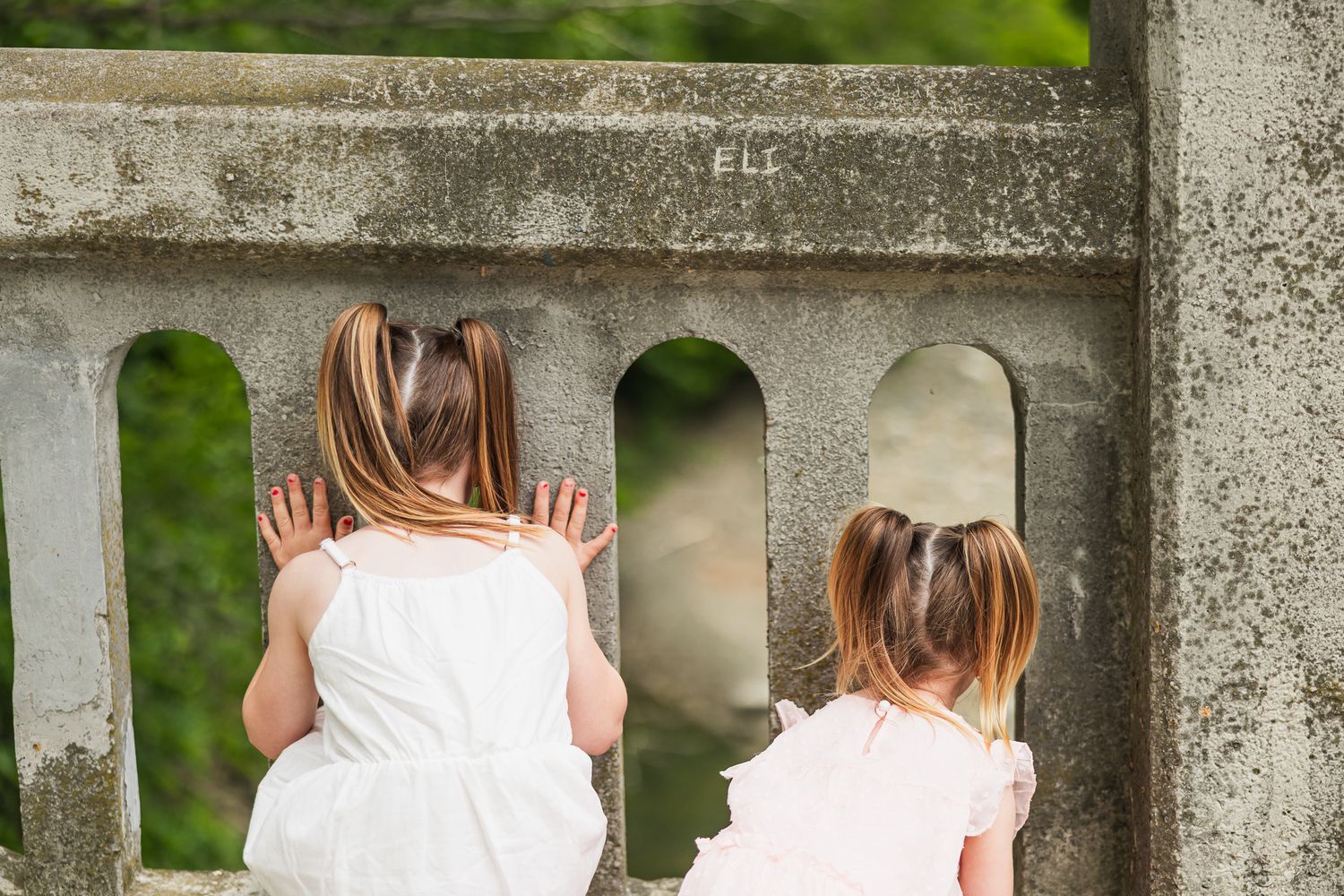 Two children in white dresses looking through stone balcony pillars in an outdoor garden setting.