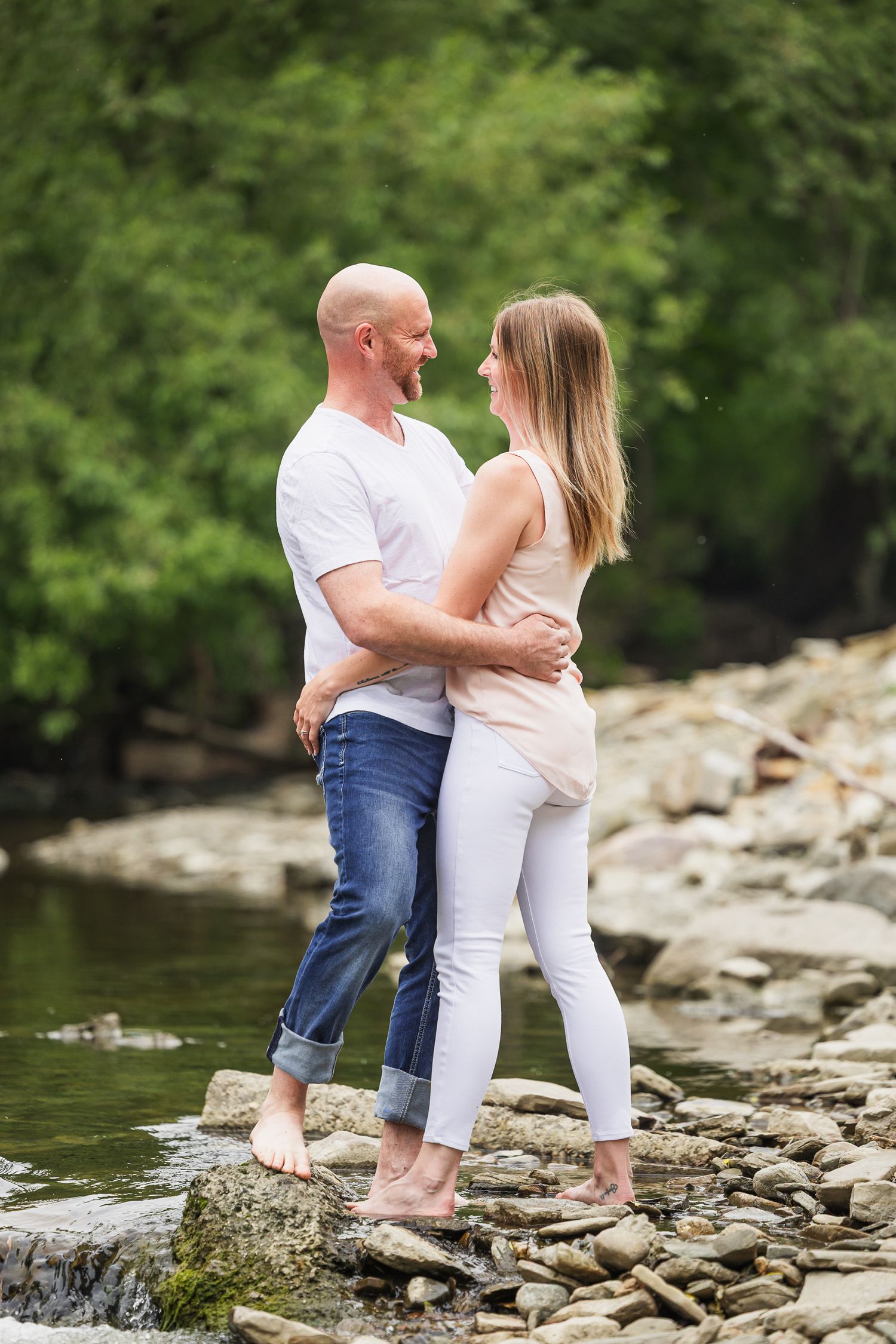 Couple sharing an intimate moment while standing barefoot on rocks by a peaceful stream.