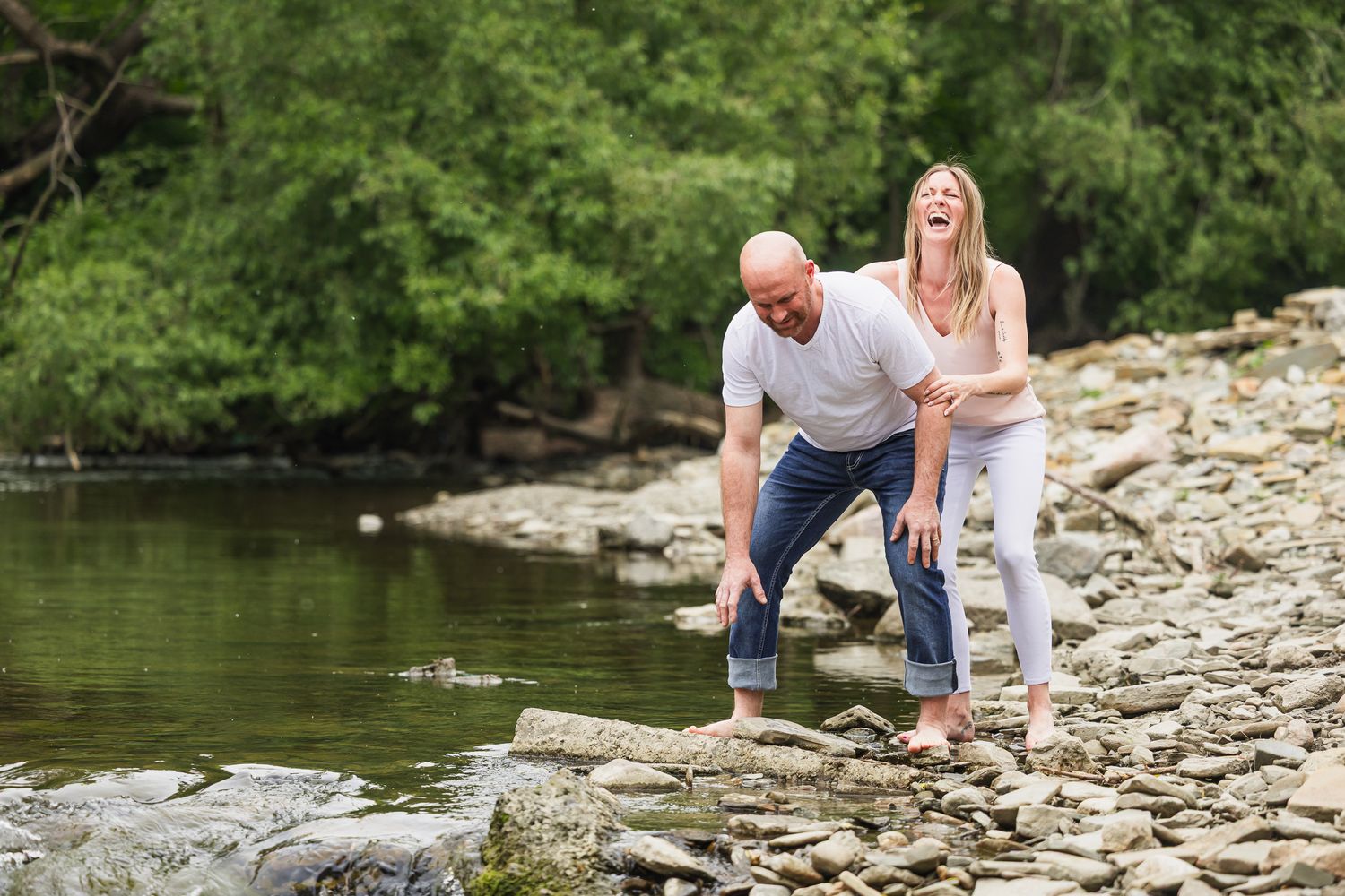 A couple sits together on rocky riverbank while taking engagement photos near flowing water.