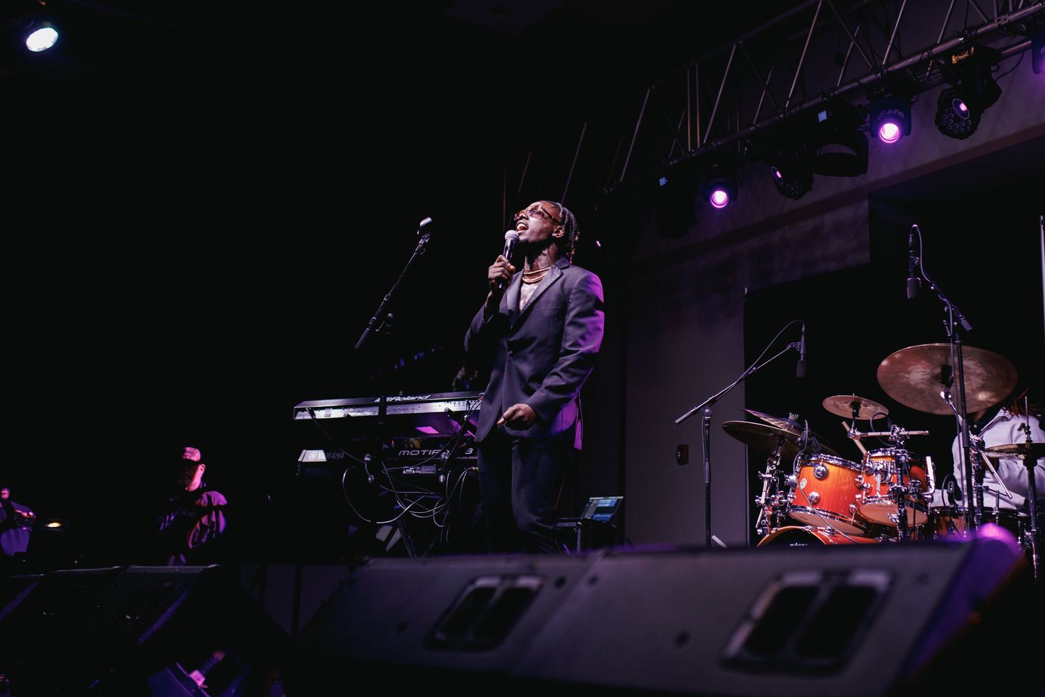 Singer performing on stage at a live concert with keyboard and drum set visible under purple stage lighting.