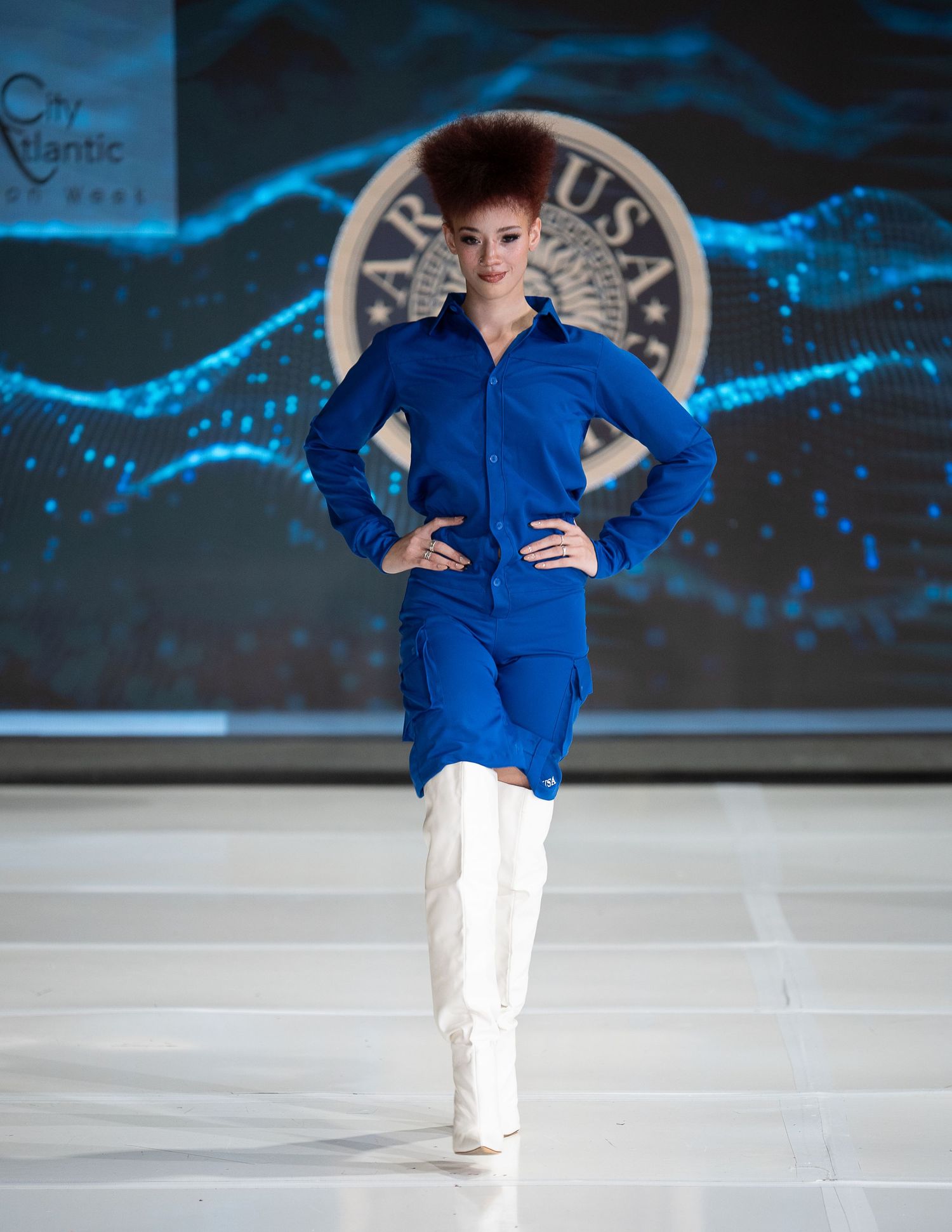 Model walking runway in bright blue fitted jumpsuit with white boots against illuminated backdrop with circular logo.