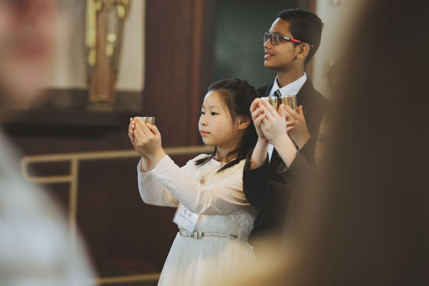 Two people join hands in prayer during a church service, one wearing white dress and another in dark attire.
