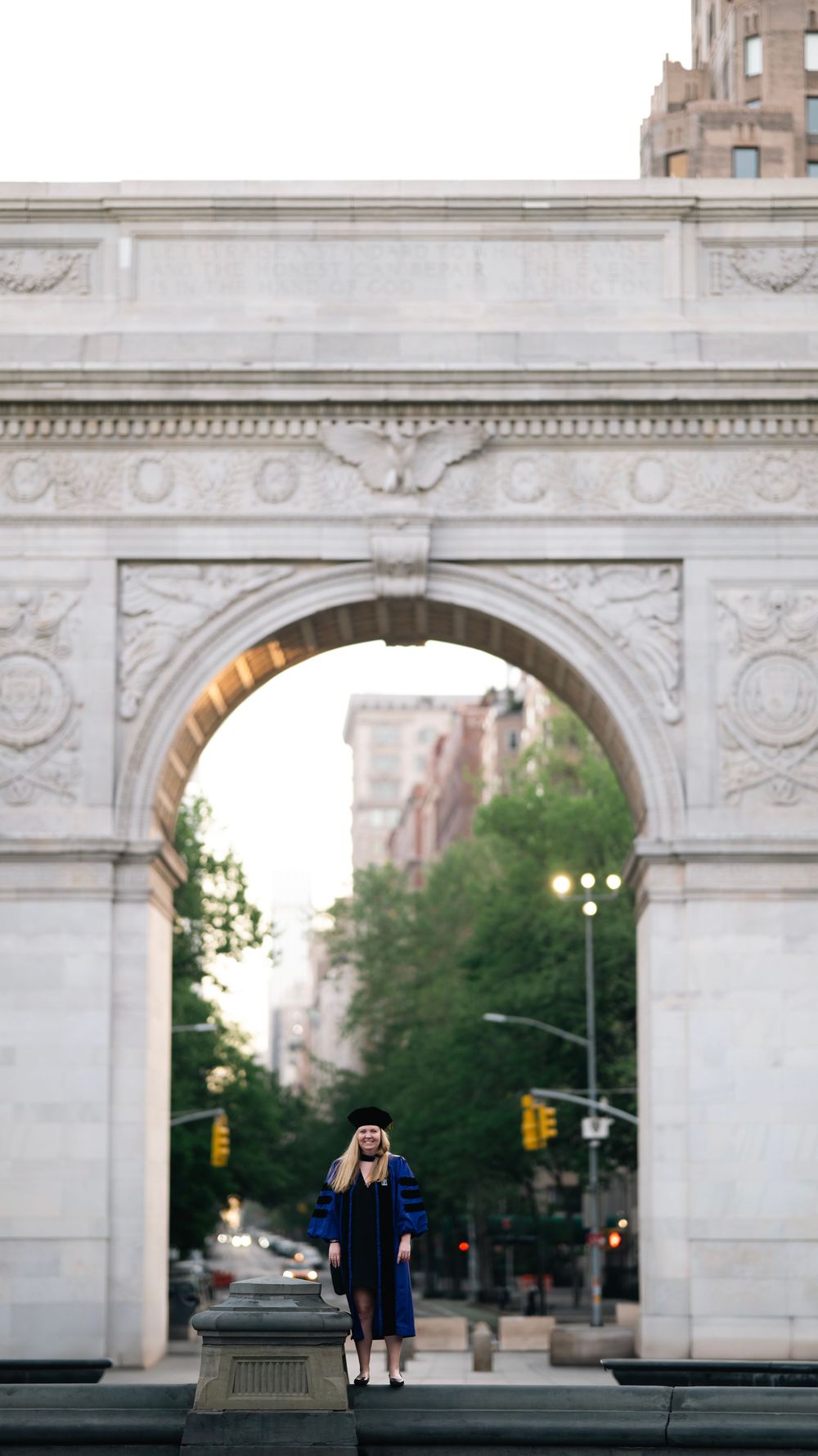 Figure in graduation gown stands beneath iconic Washington Square Arch in Manhattan at twilight hour.