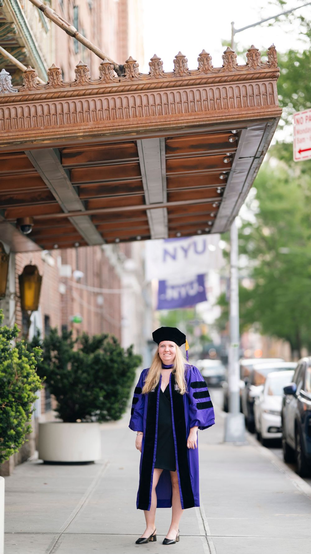 Graduate in blue and black academic regalia stands beneath ornate architectural details with NYU flag visible in background.
