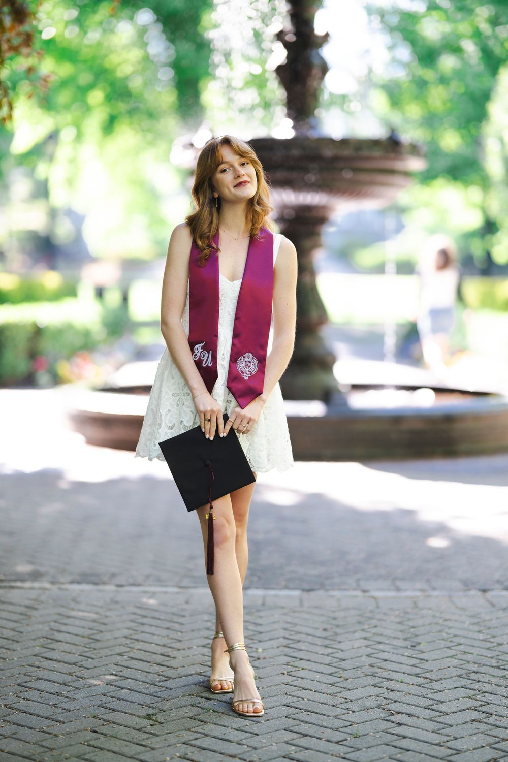 Graduate in white dress and burgundy graduation stole poses cheerfully in front of outdoor fountain on sunny day.