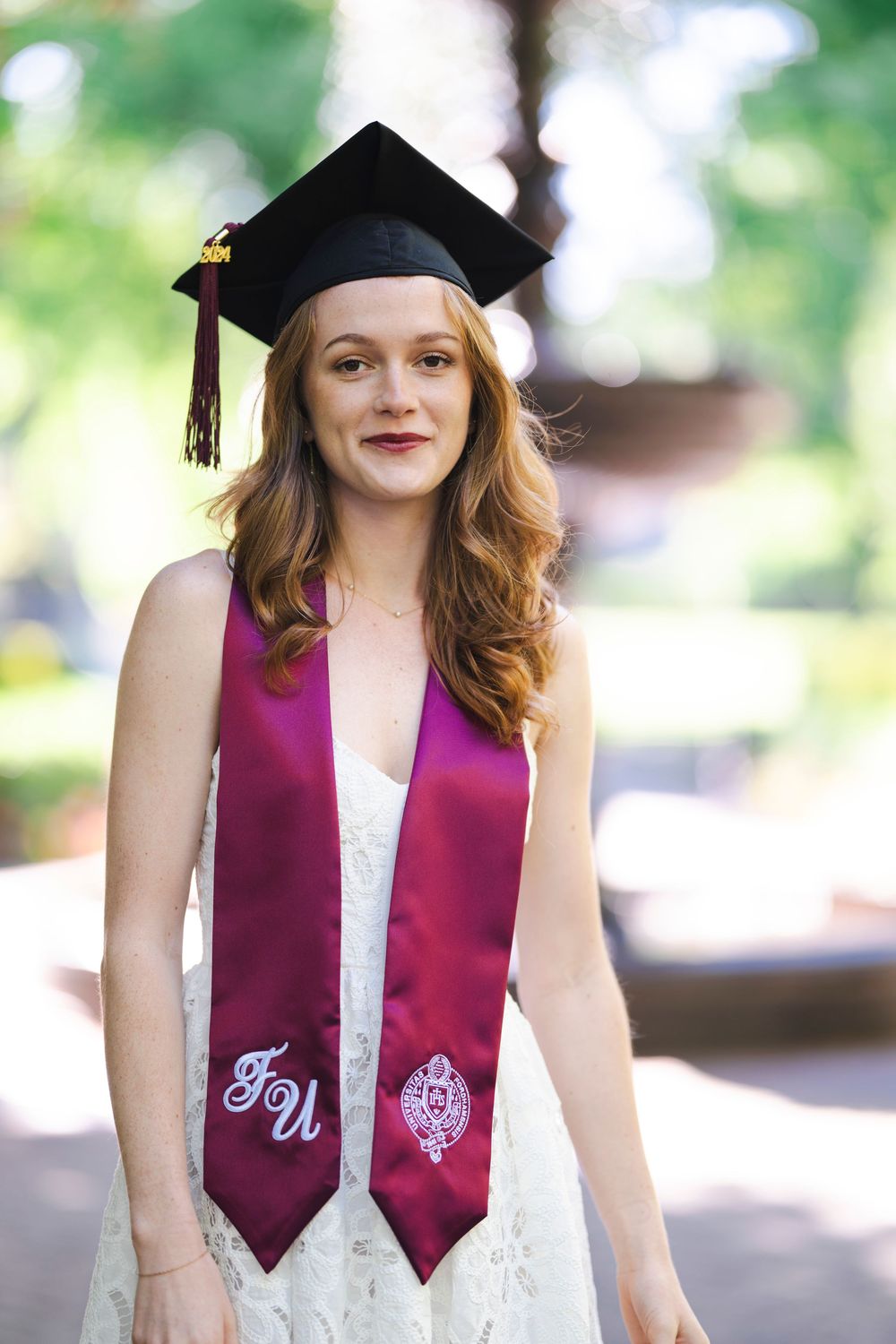 Graduate wearing burgundy graduation stole with FU letters and black mortarboard cap stands outdoors in white lace dress.