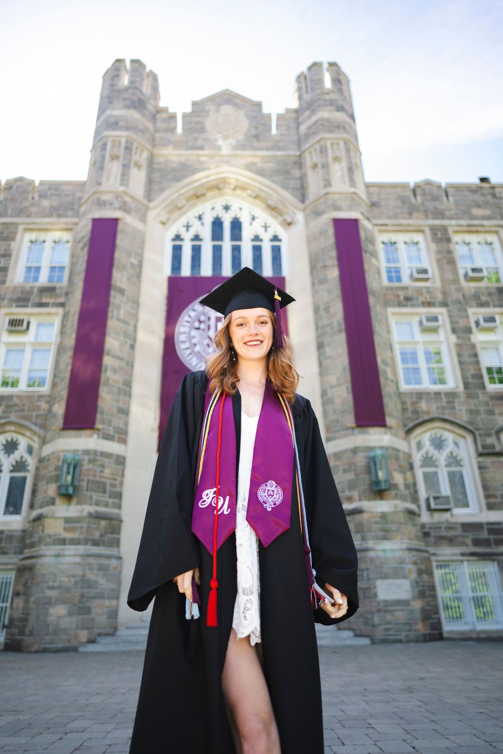 Graduate in black cap and gown with purple stoles standing in front of gothic university building architecture.