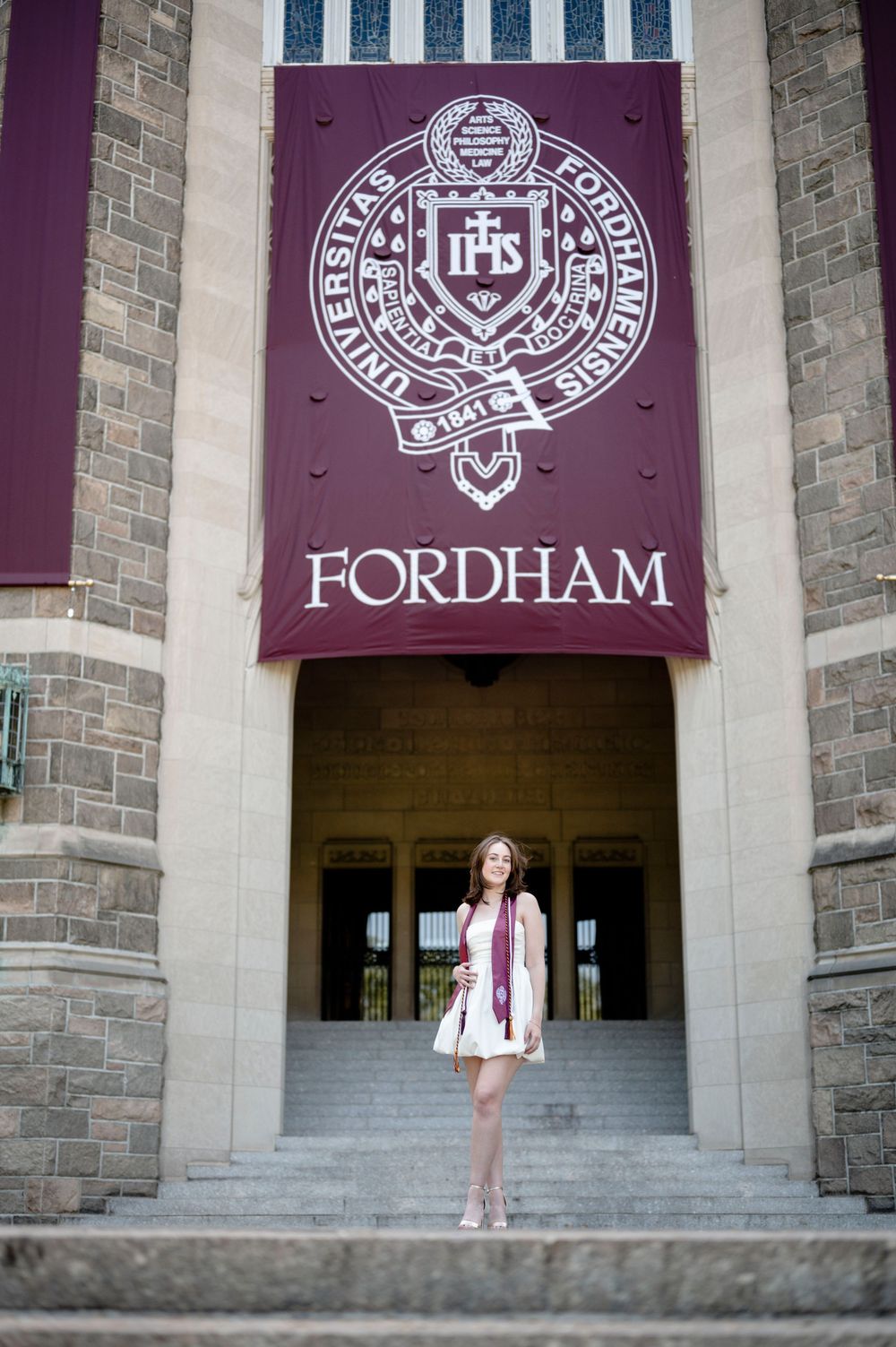 A collegiate banner displays the Fordham University seal and logo above stone entrance steps to a campus building.