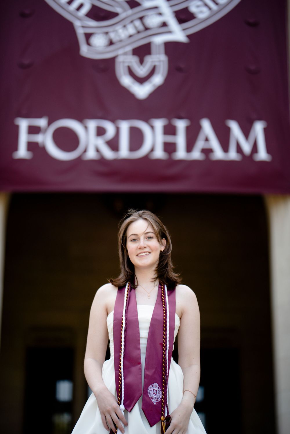 Graduate poses beneath Fordham University sign wearing graduation stole and white dress.