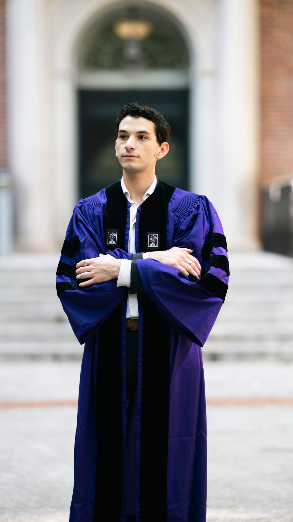 Graduate stands confidently in blue and black academic regalia with arms crossed outside a brick building entrance.