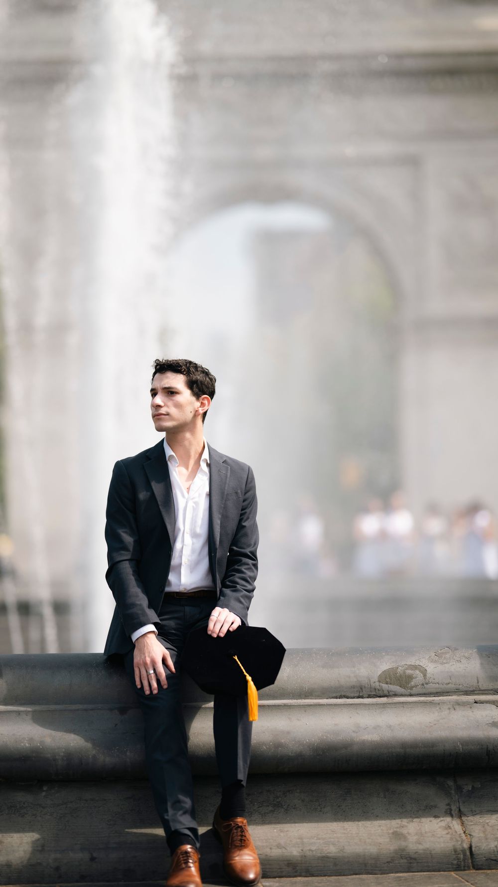 A person in a suit sits on stone steps with a graduation cap nearby and an elegant archway in the soft focus background.