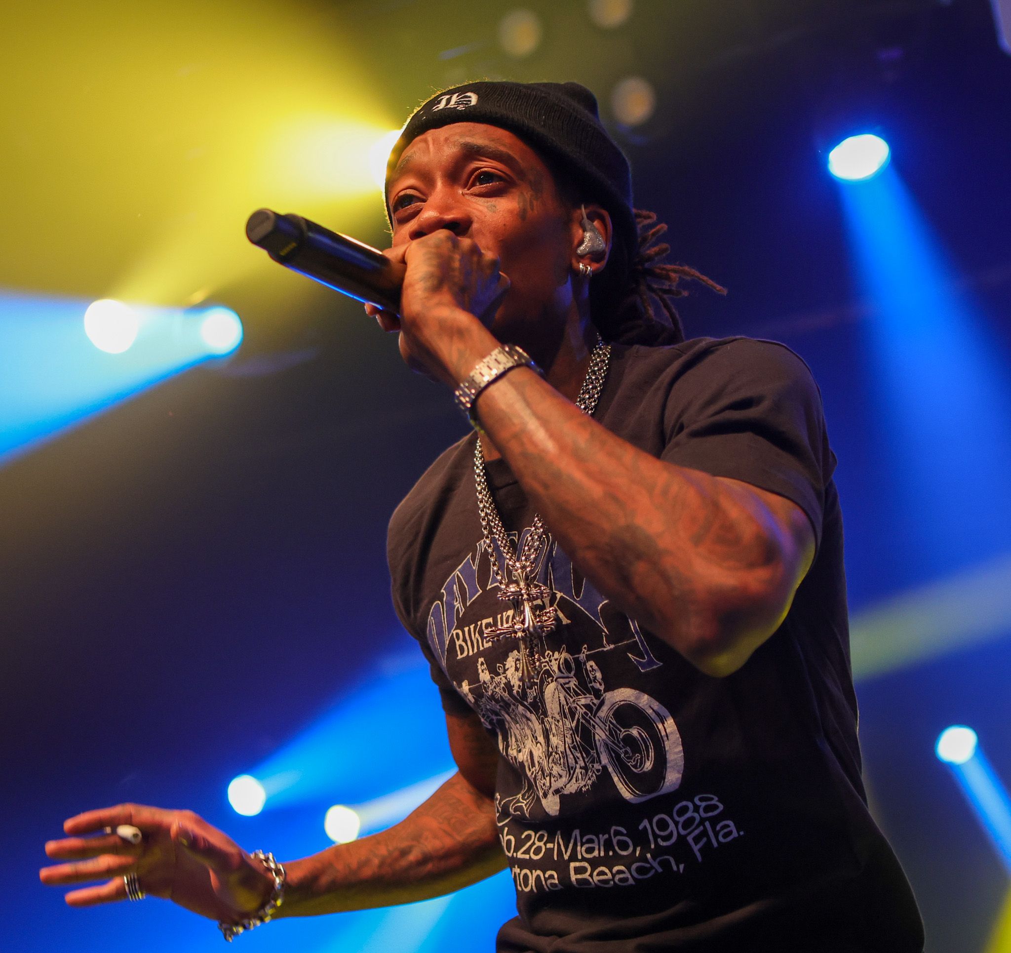 A male performer sings into a microphone on stage, wearing a black t-shirt and chain necklace under colorful concert lights.